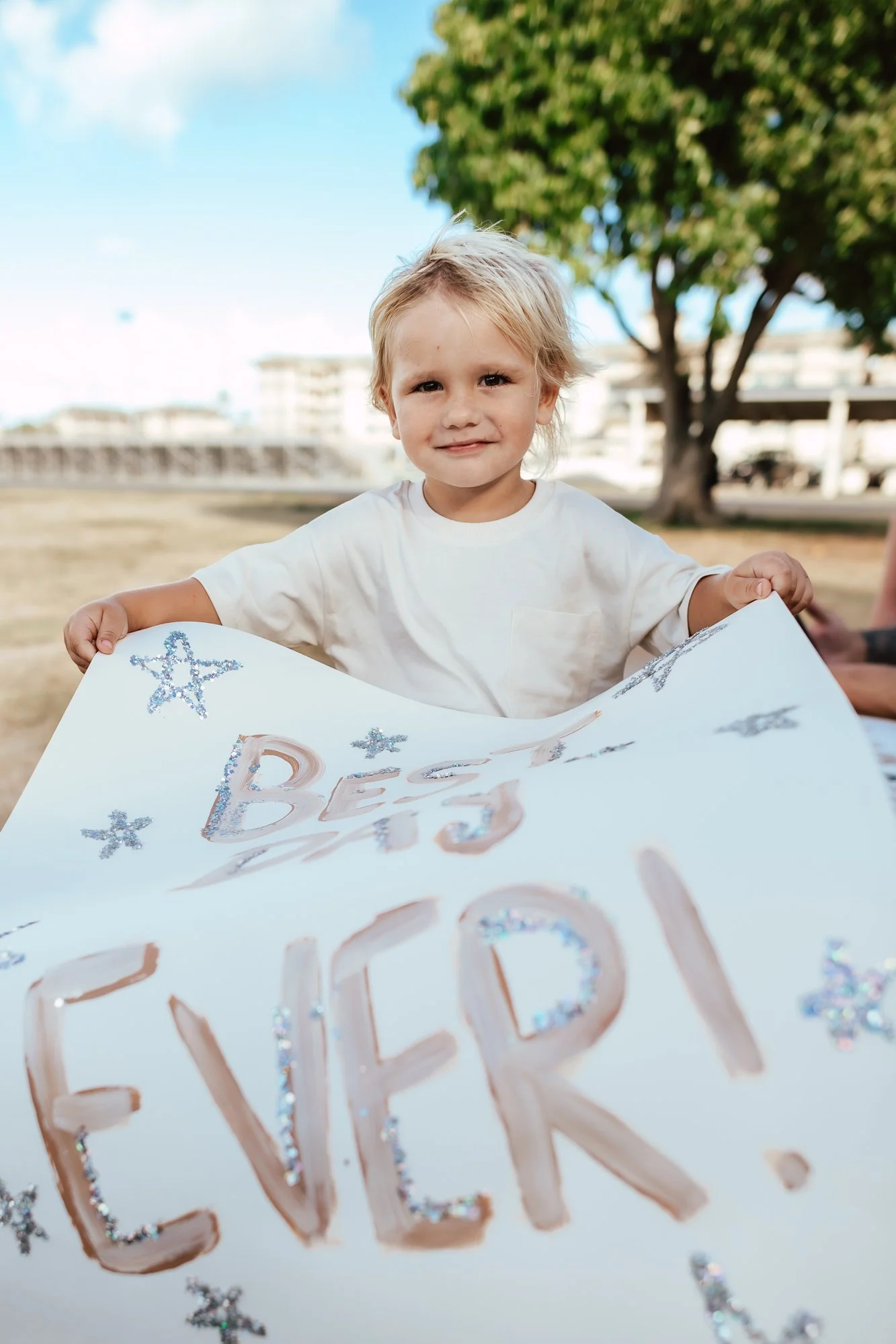 A young boy smiling outdoors, holding a large decorated sign that reads 'BIG BEACH!' with star-shaped glitter decorations. San Antonio Military Homecoming Photographer