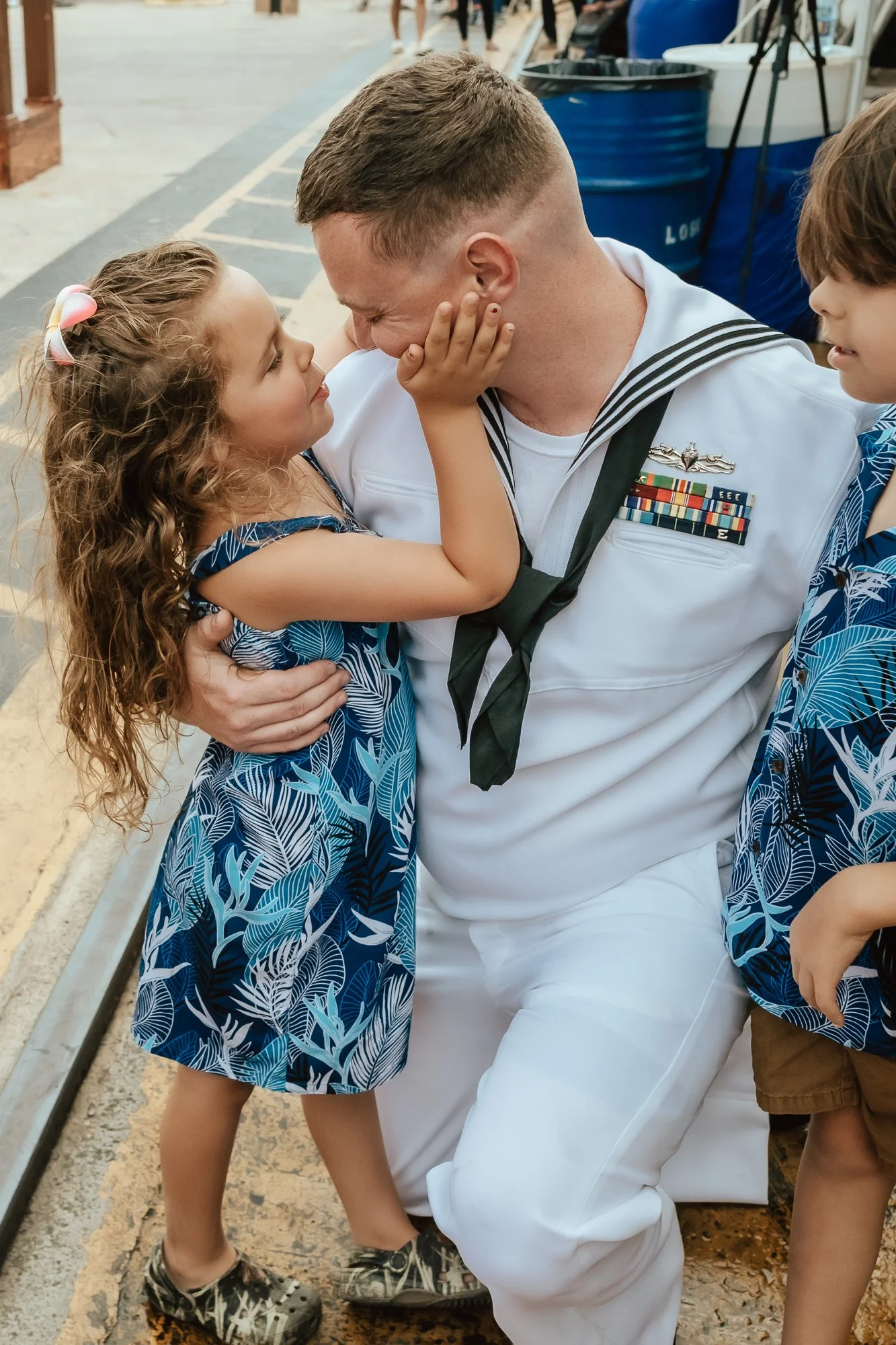A sailor in uniform is kneeling, embracing a young girl who is touching his face and looking into his eyes, with another child nearby. They are outdoors near a street or parade route. San Antonio Military Homecoming Photographer