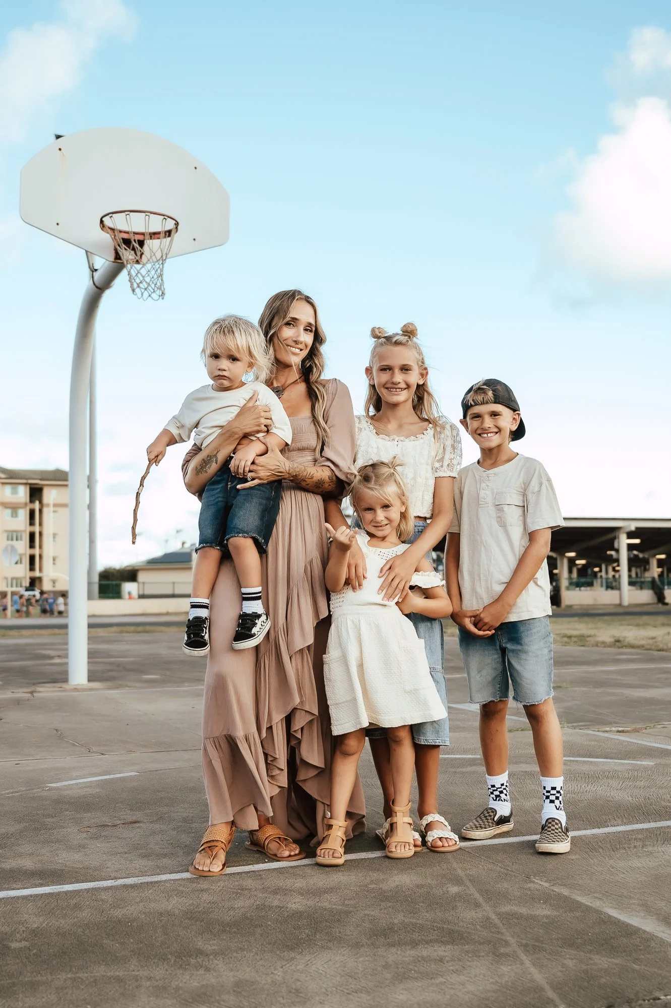 A group of five people, including an adult woman and four children, smiling and standing together outside on a basketball court during daytime. There is a basketball hoop in the background. San Antonio Military Homecoming Photographer