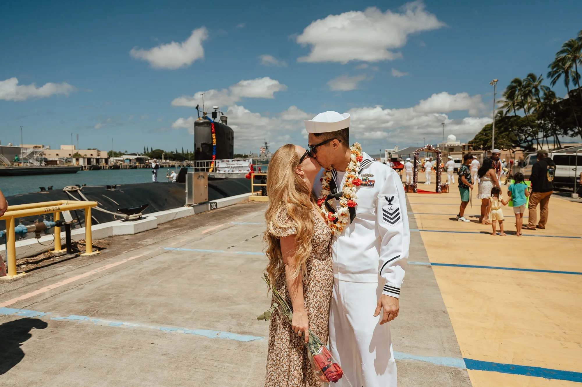 A couple in a wedding ceremony at a dock, with a woman in a floral dress holding roses and a man in a navy sailor uniform, sharing a kiss surrounded by people and sailor ships under a partly cloudy sky. San Antonio Military Homecoming Photographer