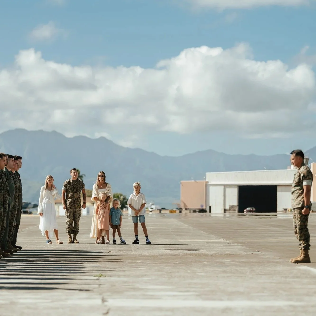 Military personnel and civilians standing on an airstrip, with mountains and a hangar in the background, in a formal gathering or ceremony. San Antonio Military Homecoming Photographer