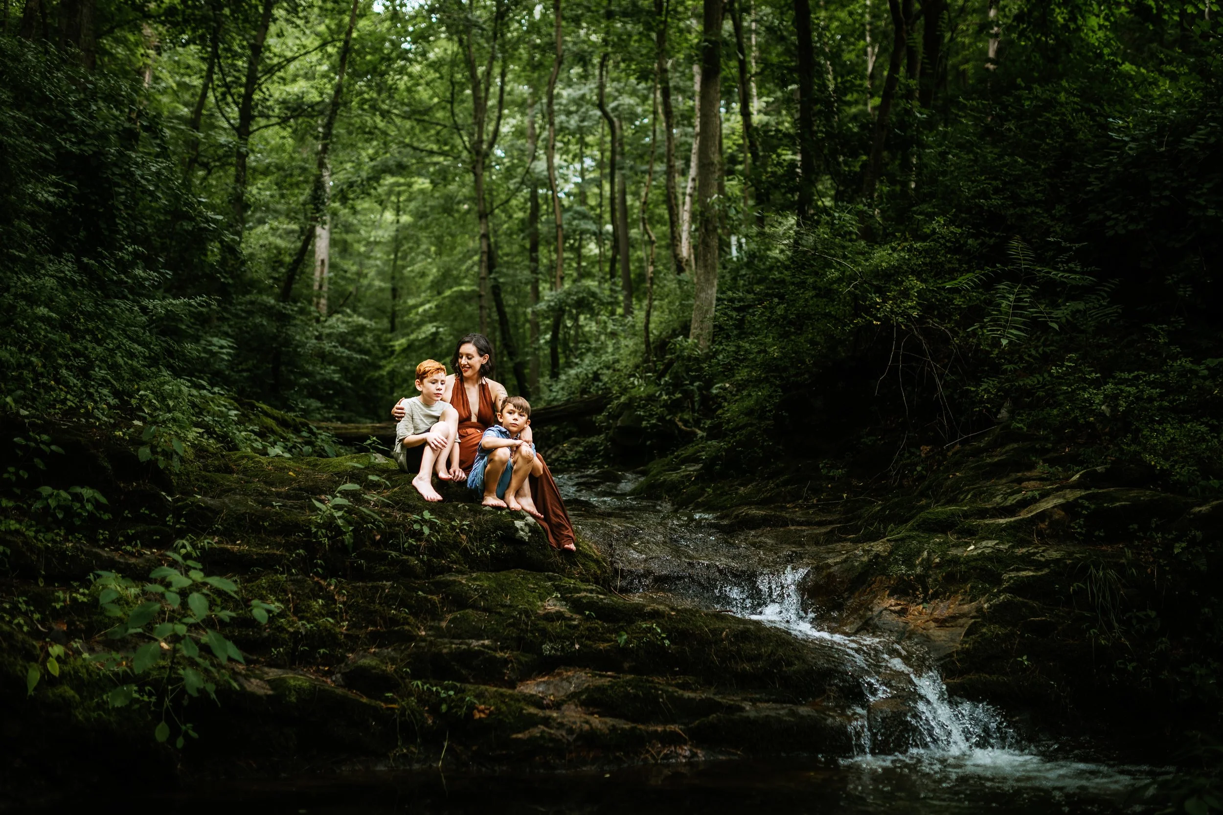 A woman and two young boys sitting on mossy rocks beside a small creek in a dense green forest. San Antonio Family and Birth Photographer
