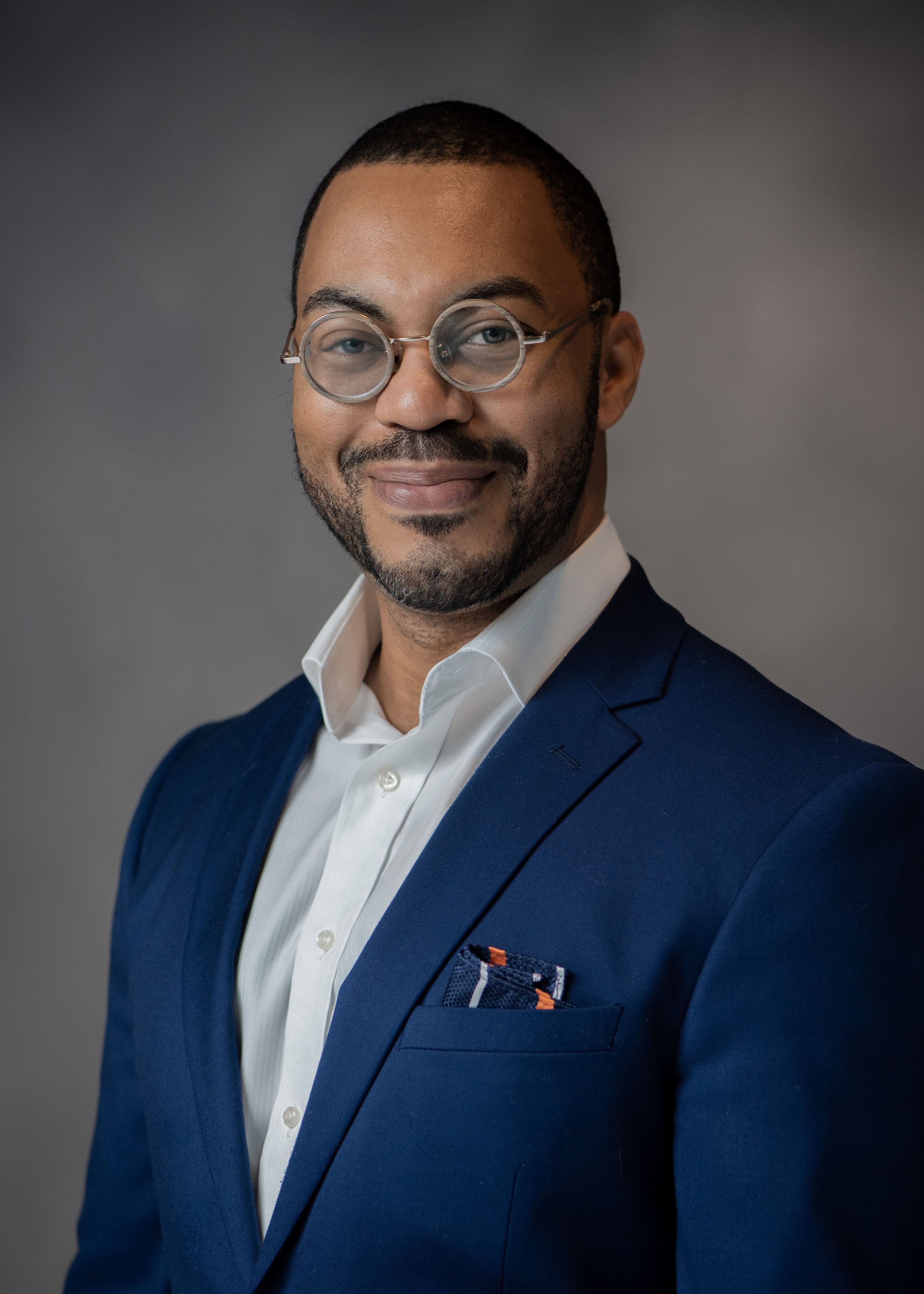 portrait of Renaldo smiling. He is wearing a white shirt, blue suit jacket, and brown circular glasses.