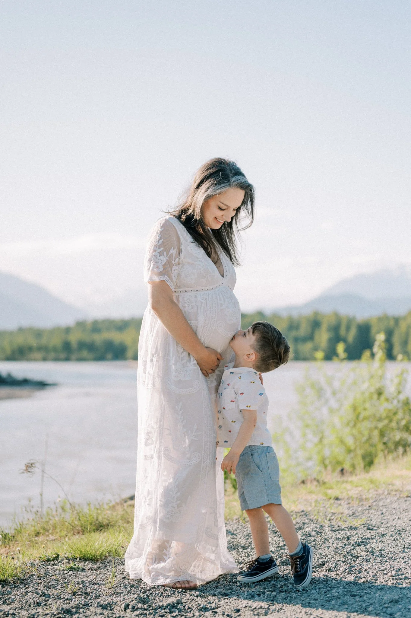 A pregnant woman in a white lace dress stands outdoors by a river, with a small boy kissing her belly.