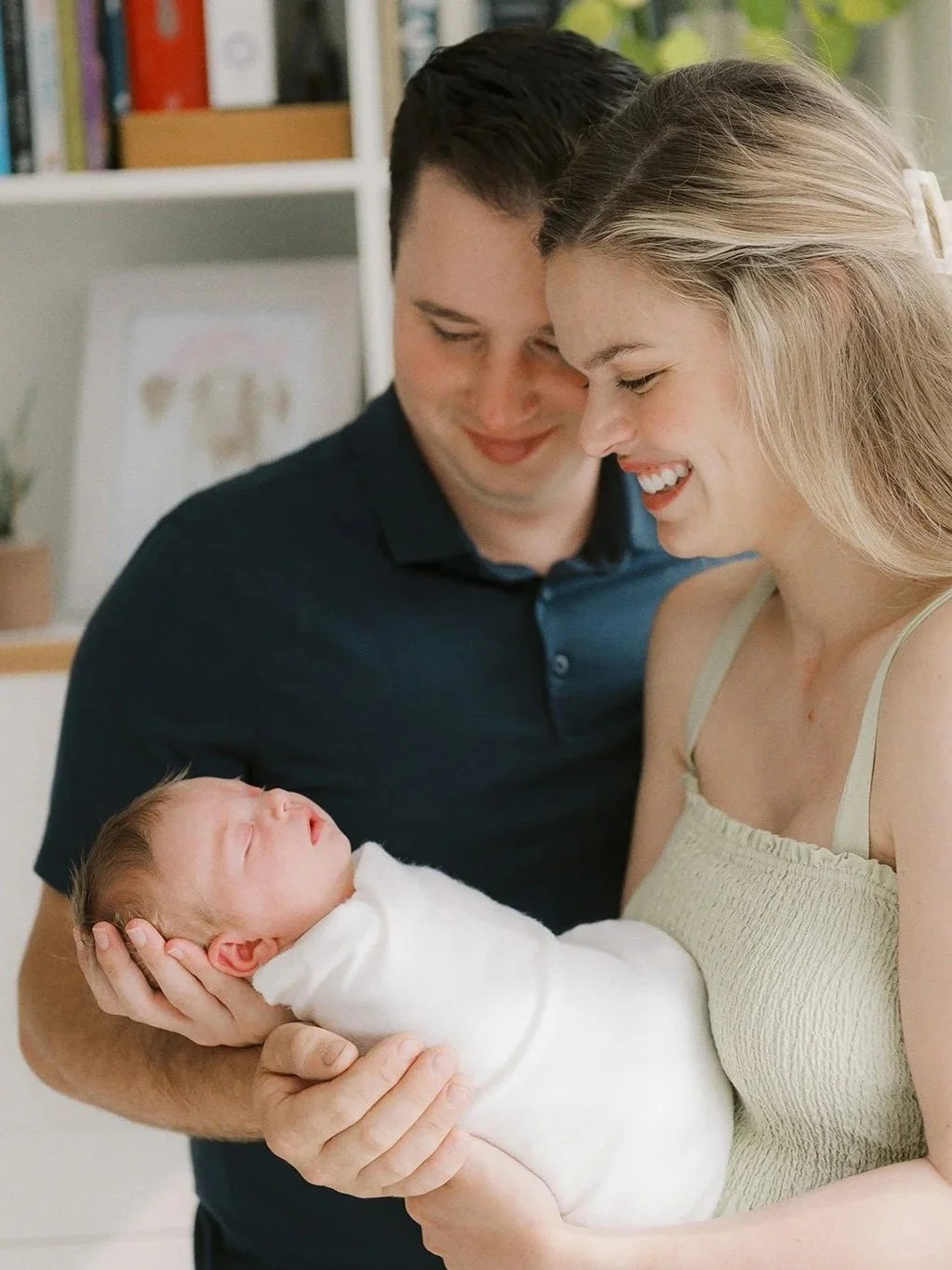 A woman holding a newborn baby, with a man standing beside them, both smiling and looking at the baby, inside a home with books on shelves in the background.