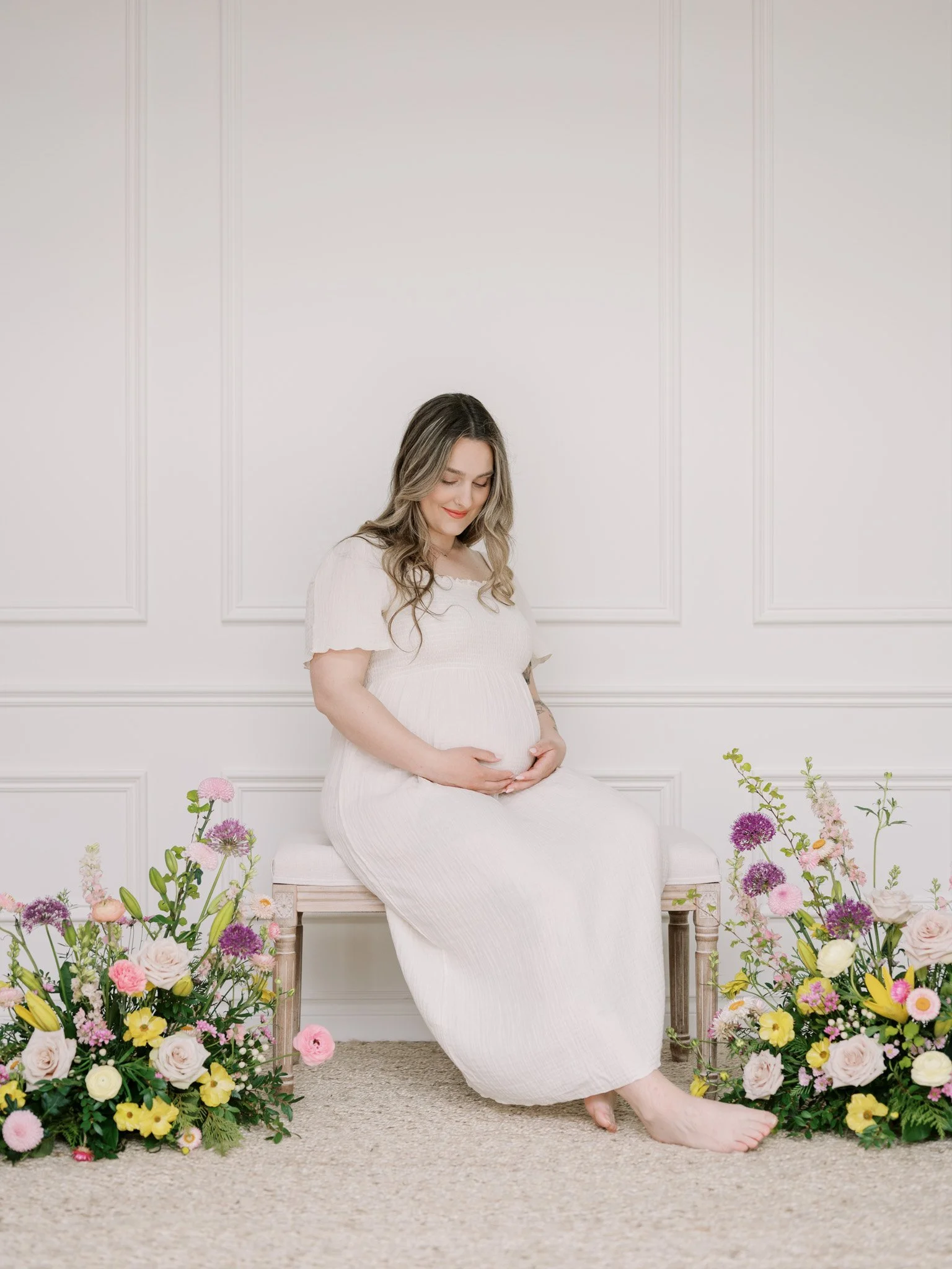 Pregnant woman in a long white dress sitting on a bench, surrounded by colorful flowers, touching her belly and smiling, against a white paneled wall.