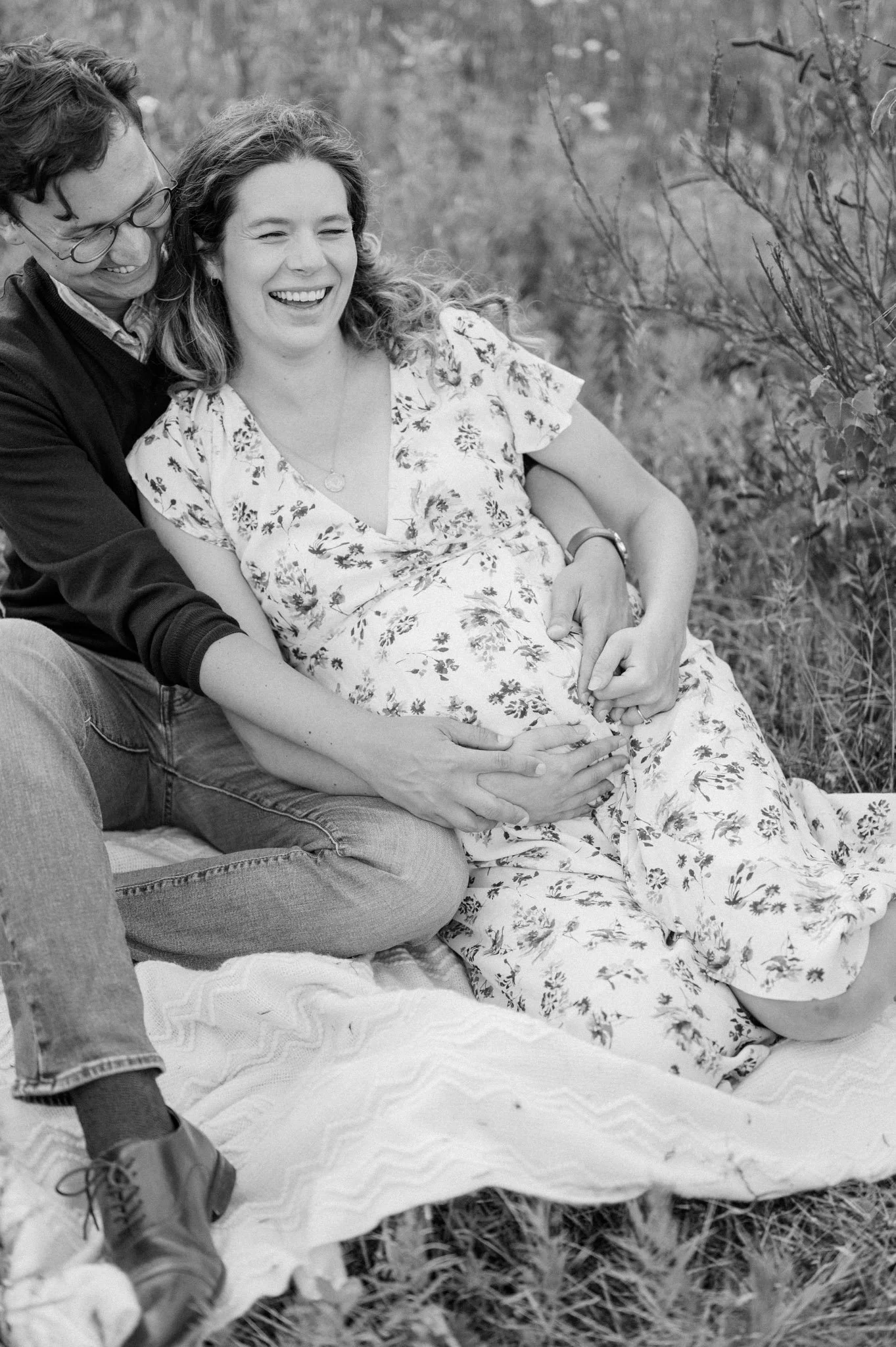 Black and white photo of a couple sitting on a blanket in a field, with the woman wearing a floral dress and her hands on her pregnant belly.