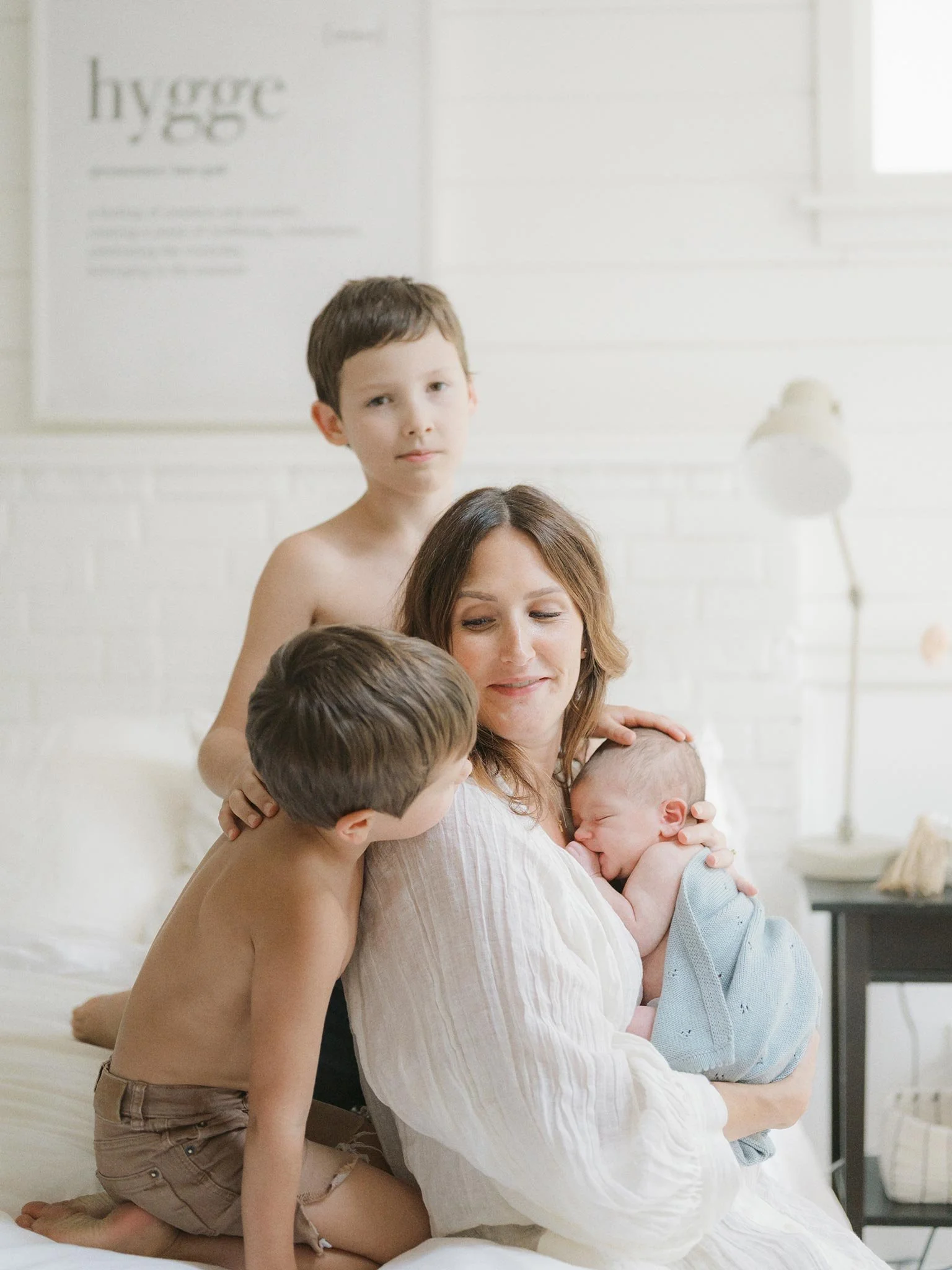 A woman holding a newborn baby, with two young boys close by, in a bright, cozy room with white walls and minimal decor.