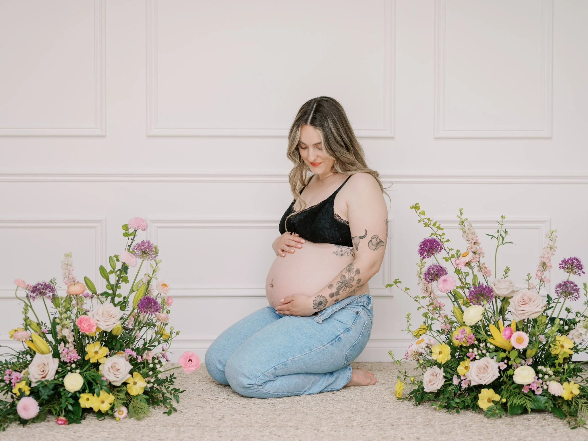 Pregnant woman in black bra and jeans kneeling between two floral arrangements against a white paneled wall.