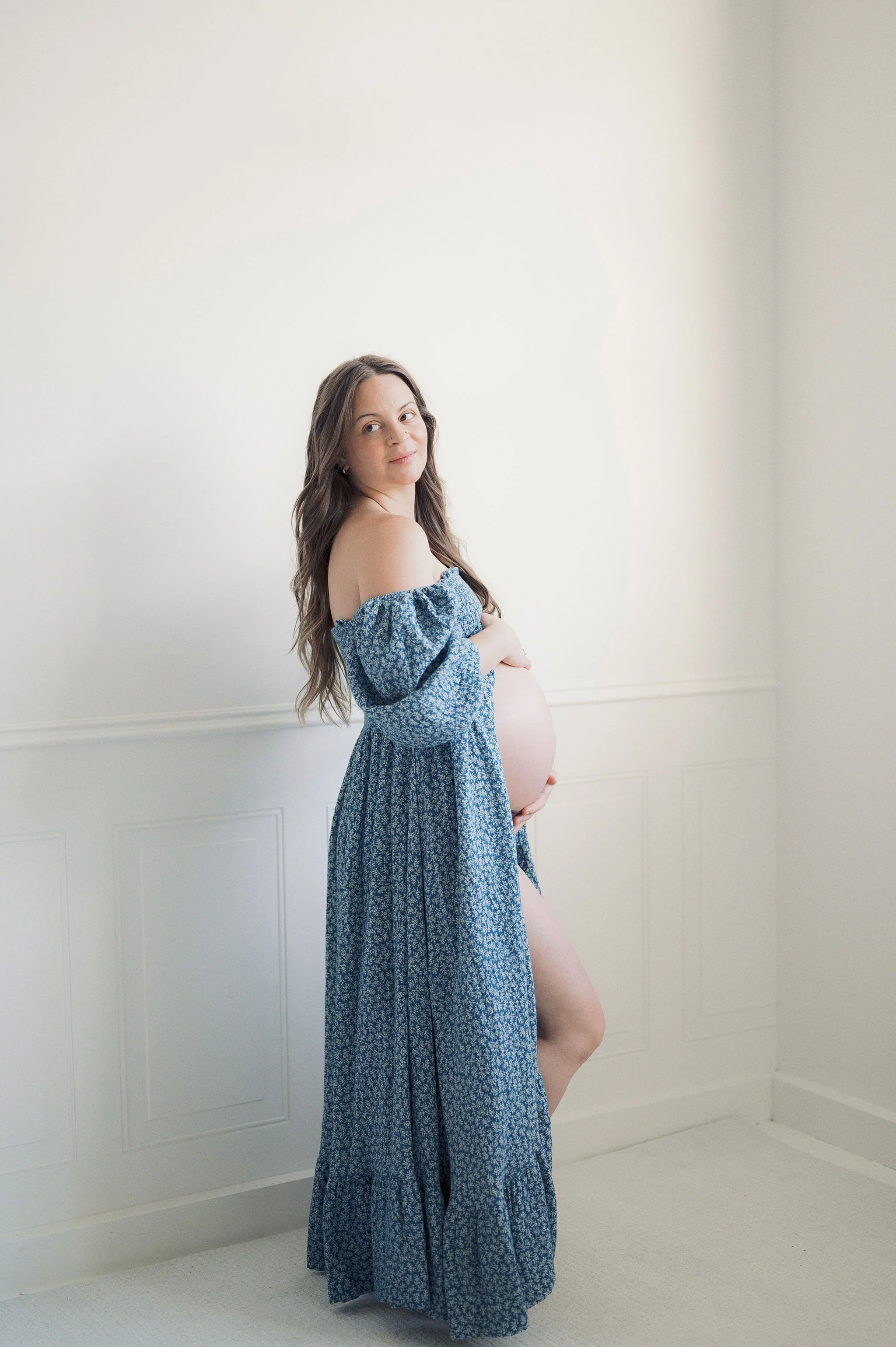 Pregnant woman in a blue dress standing against a white wall.