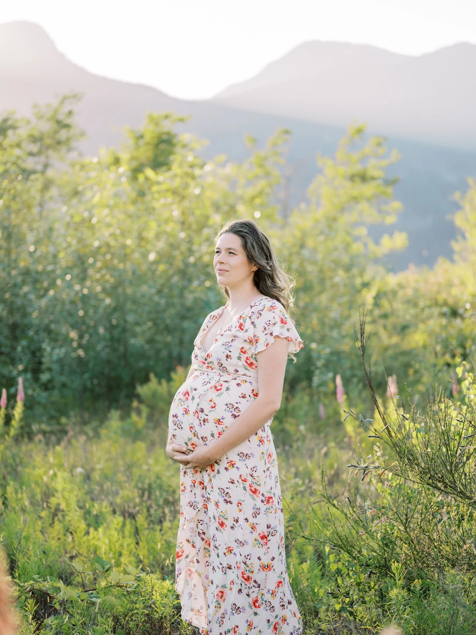 Pregnant woman in floral dress standing in a field with mountains in the background.
