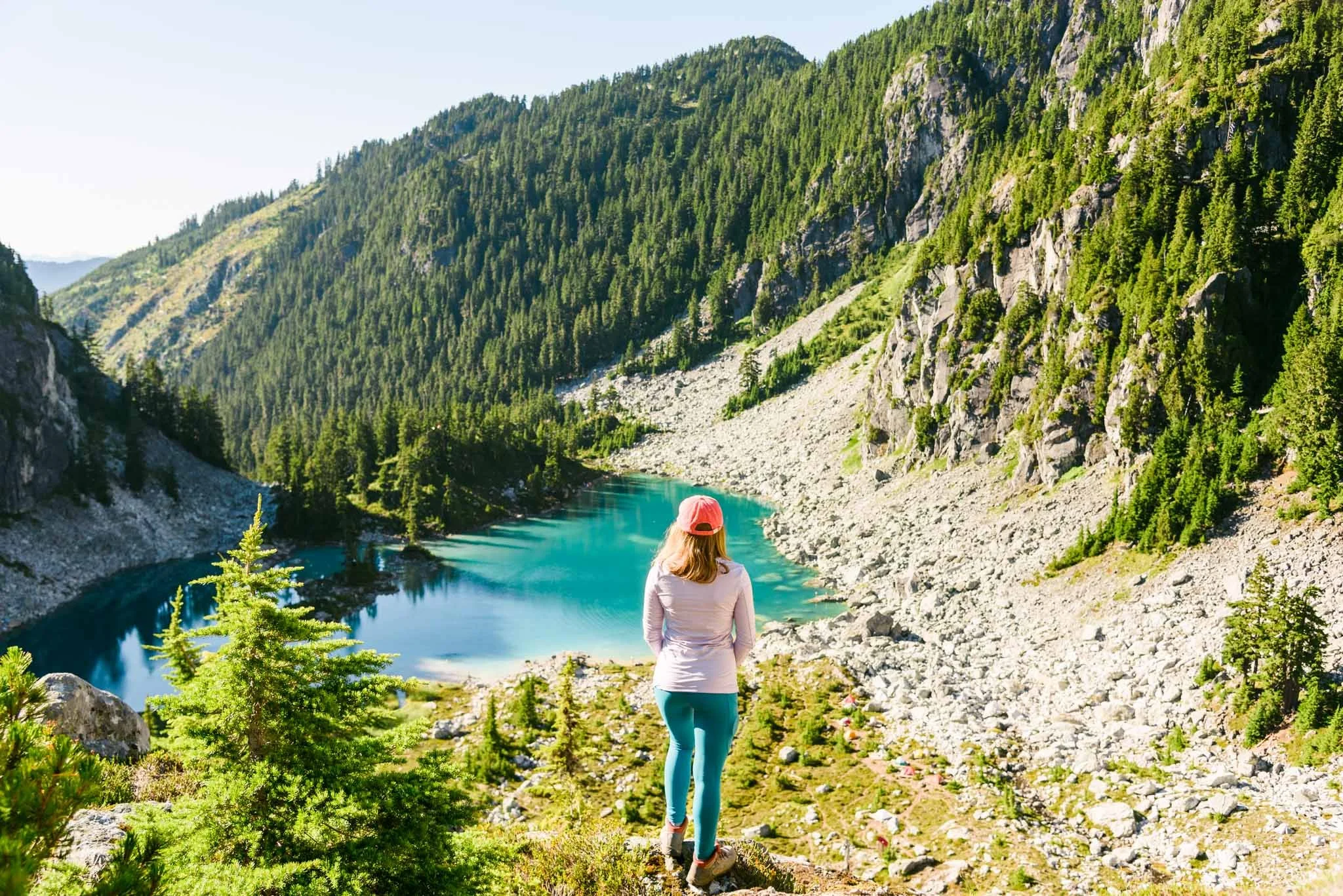 A person wearing a pink cap and outdoor clothing stands on a hill, overlooking a turquoise mountain lake surrounded by rocky terrain and dense green forest under a clear sky.