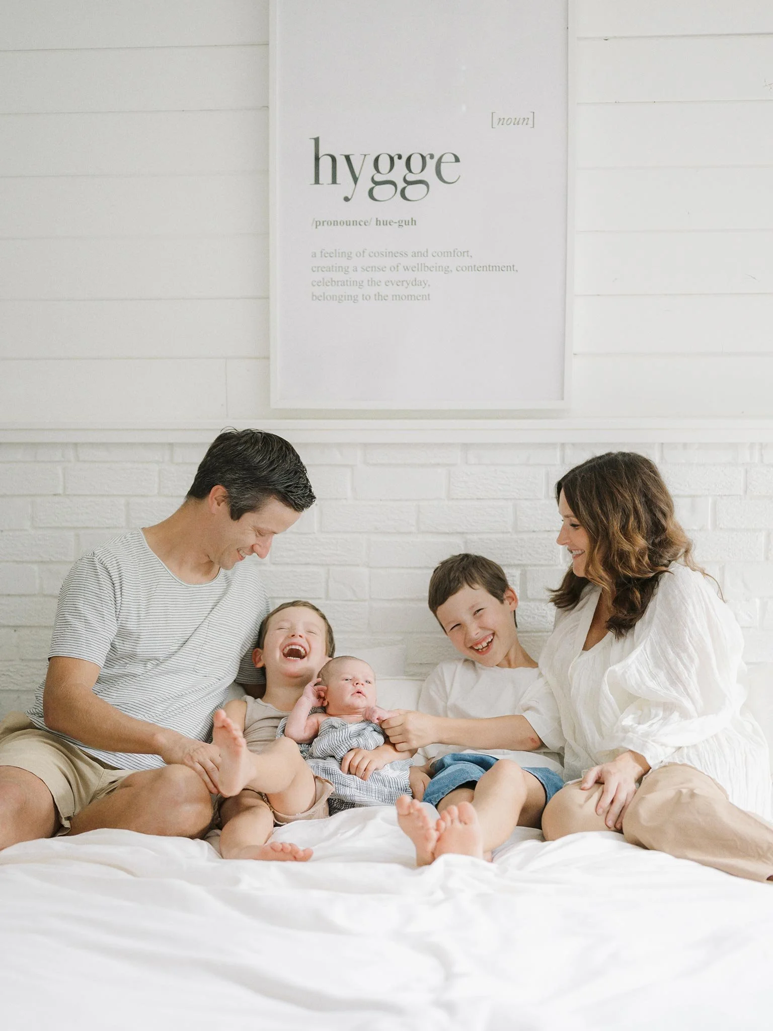 Family of five sitting on a bed, smiling and laughing. A father, mother, and three children, including a baby, enjoying a joyful moment together.