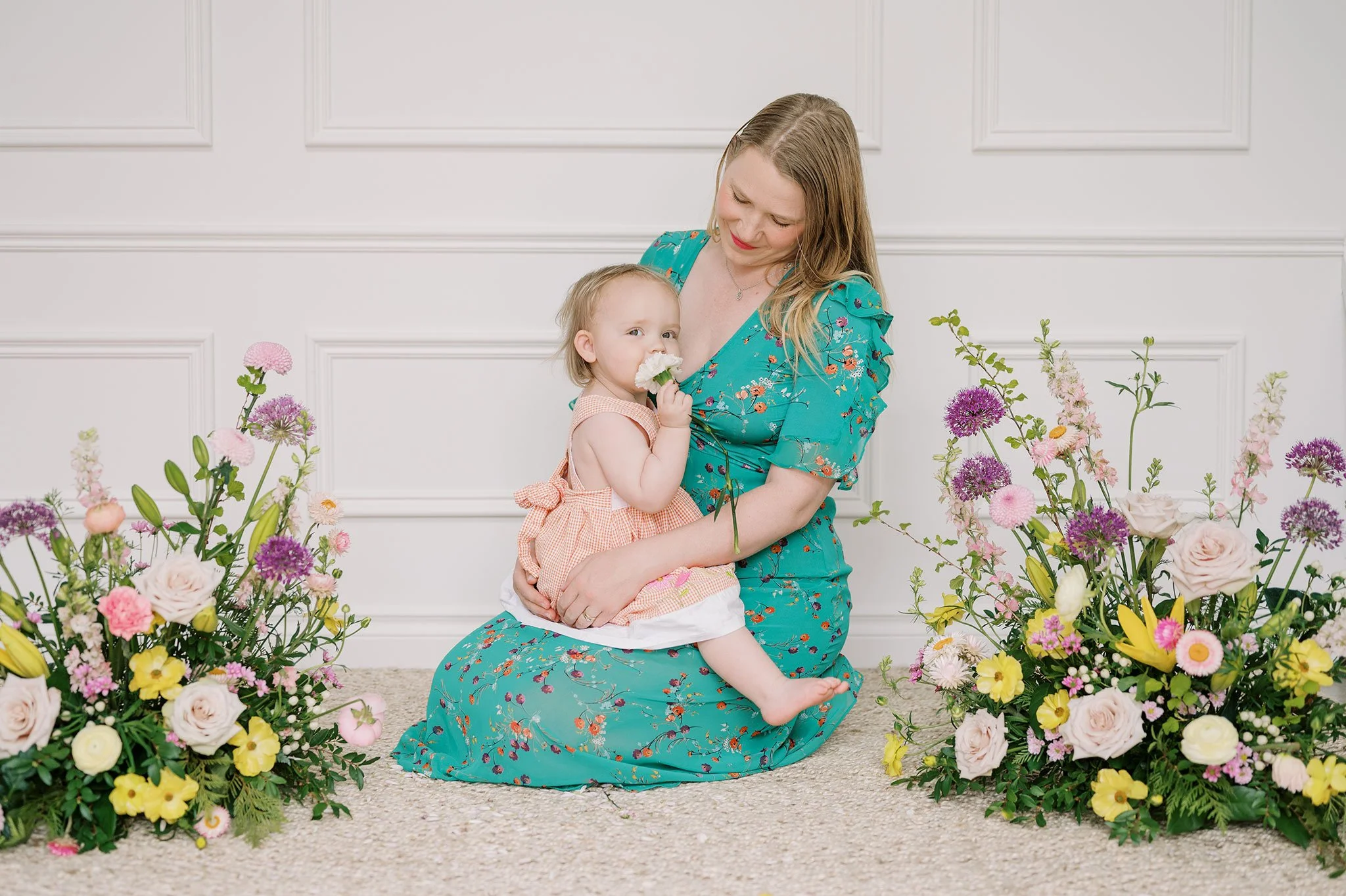 Woman in a green floral dress holding a baby surrounded by colorful flower arrangements.