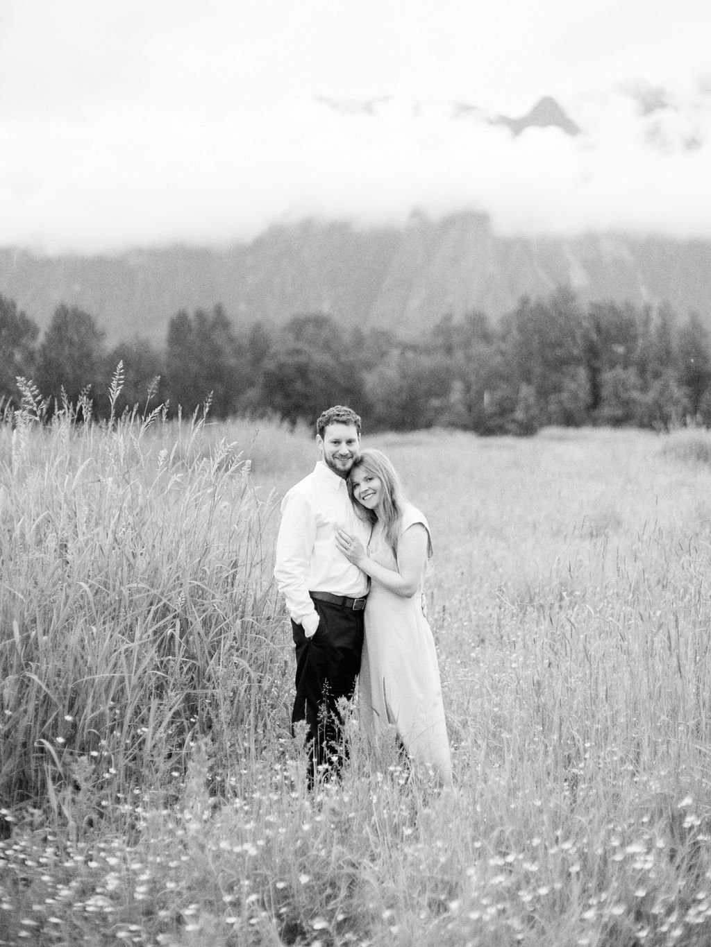 A couple standing in a grassy field with mountains and clouds in the background, captured in black and white.