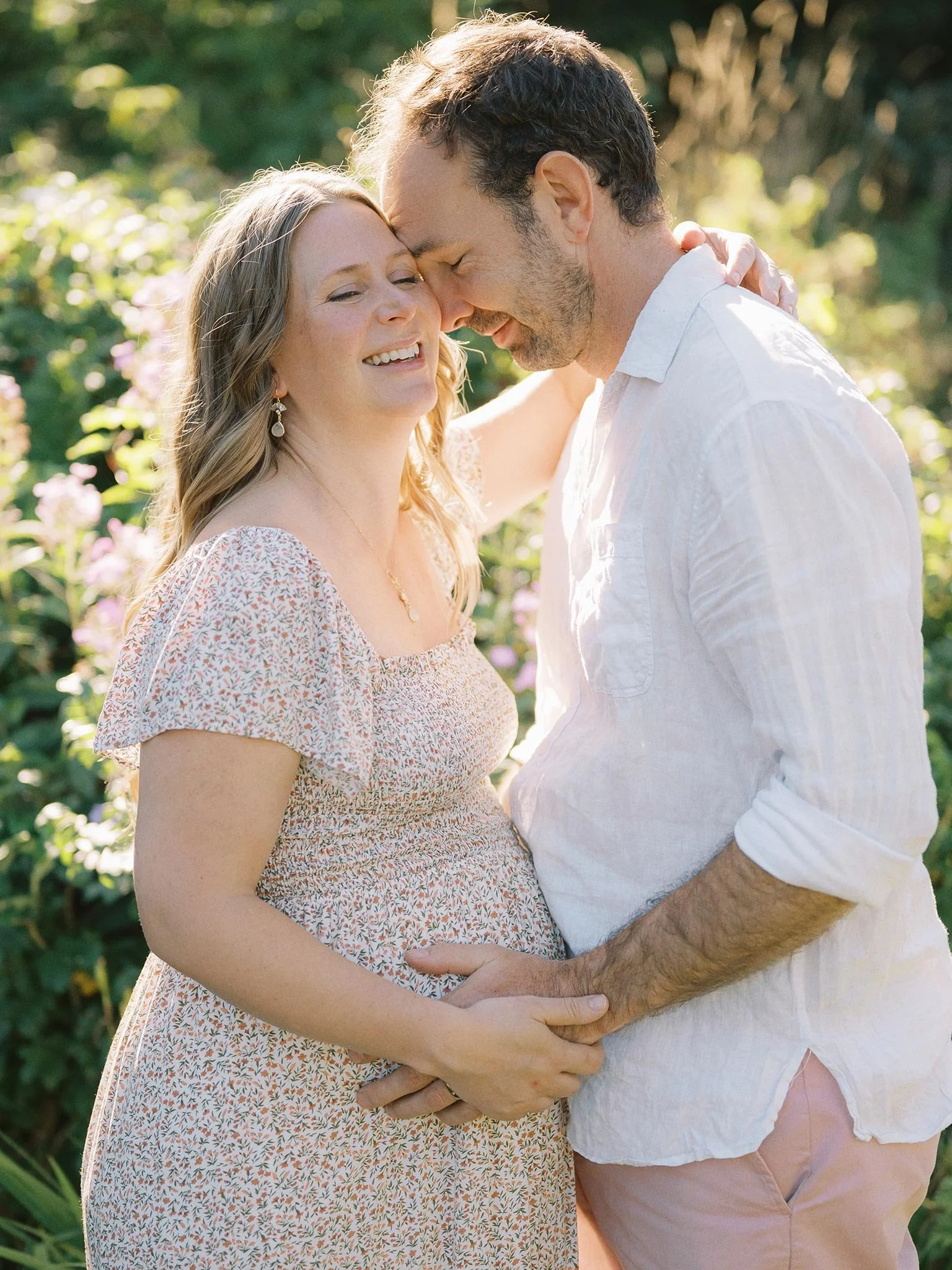 A pregnant woman and her partner smiling and touching foreheads in a garden during daylight.