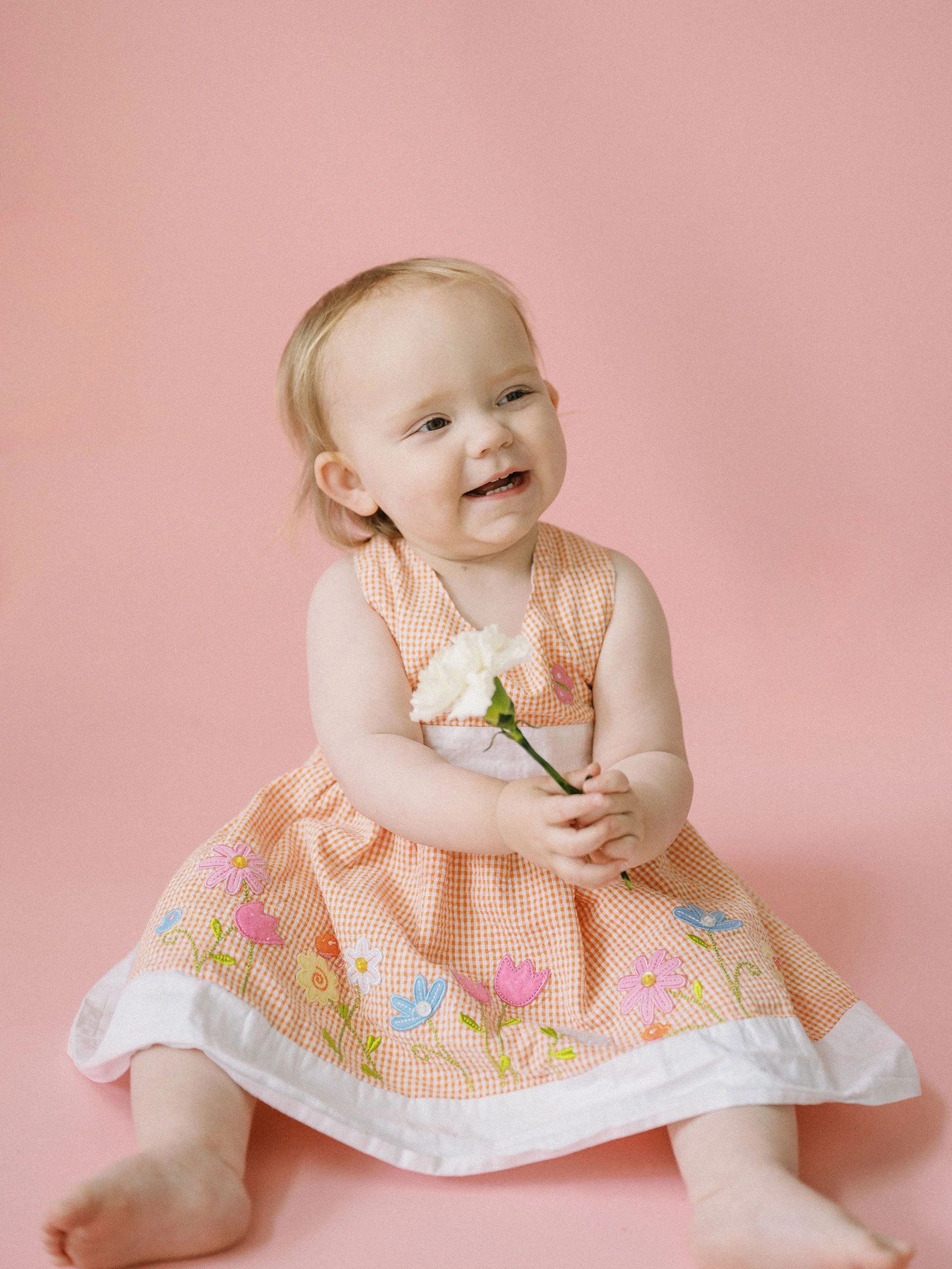 Smiling baby holding a white flower, wearing a colorful dress on a pink background.