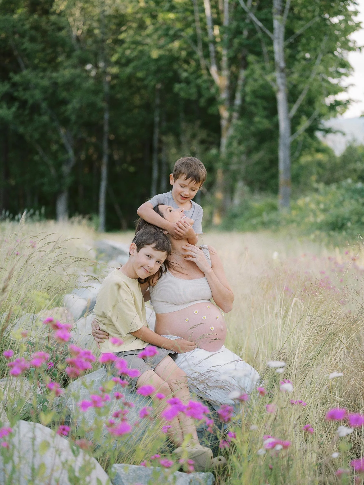 A pregnant woman sitting in a field of flowers with two young boys, one sitting next to her and the other hugging her from behind, smiling together.