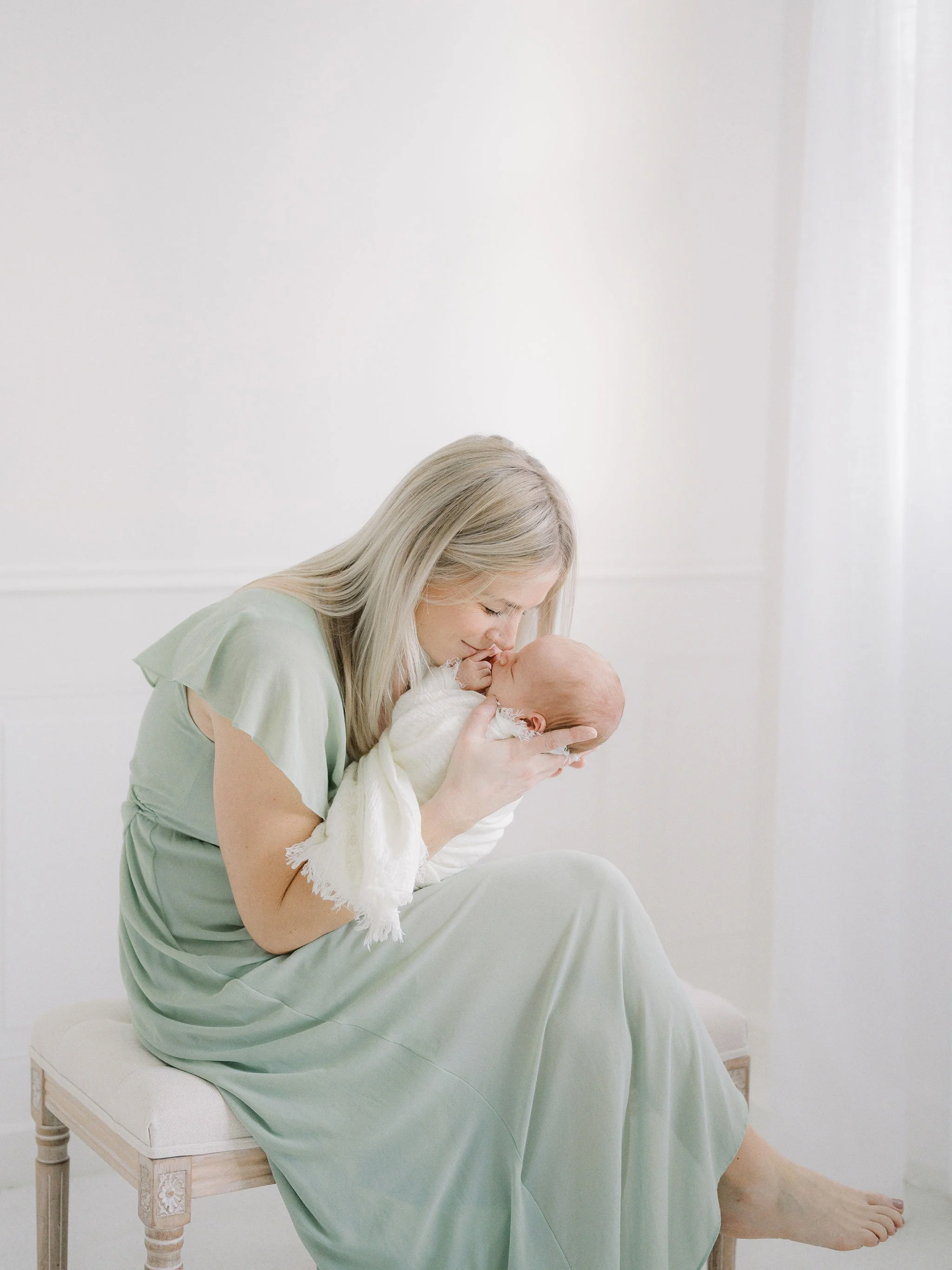 A woman in a light green dress sitting on a stool cradling a newborn baby wrapped in a white blanket.