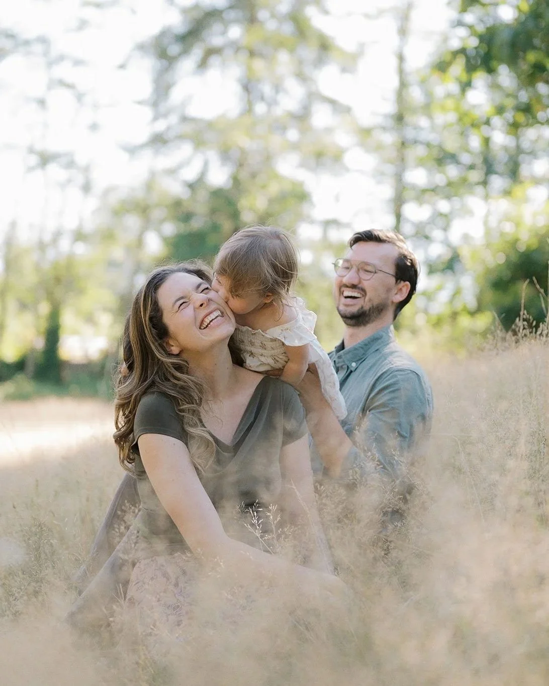 A happy family of three enjoying outdoor time in a field with tall grass, surrounded by trees, during a sunny day. The mother and father are smiling, and their young daughter is giving the mother a kiss on the cheek.