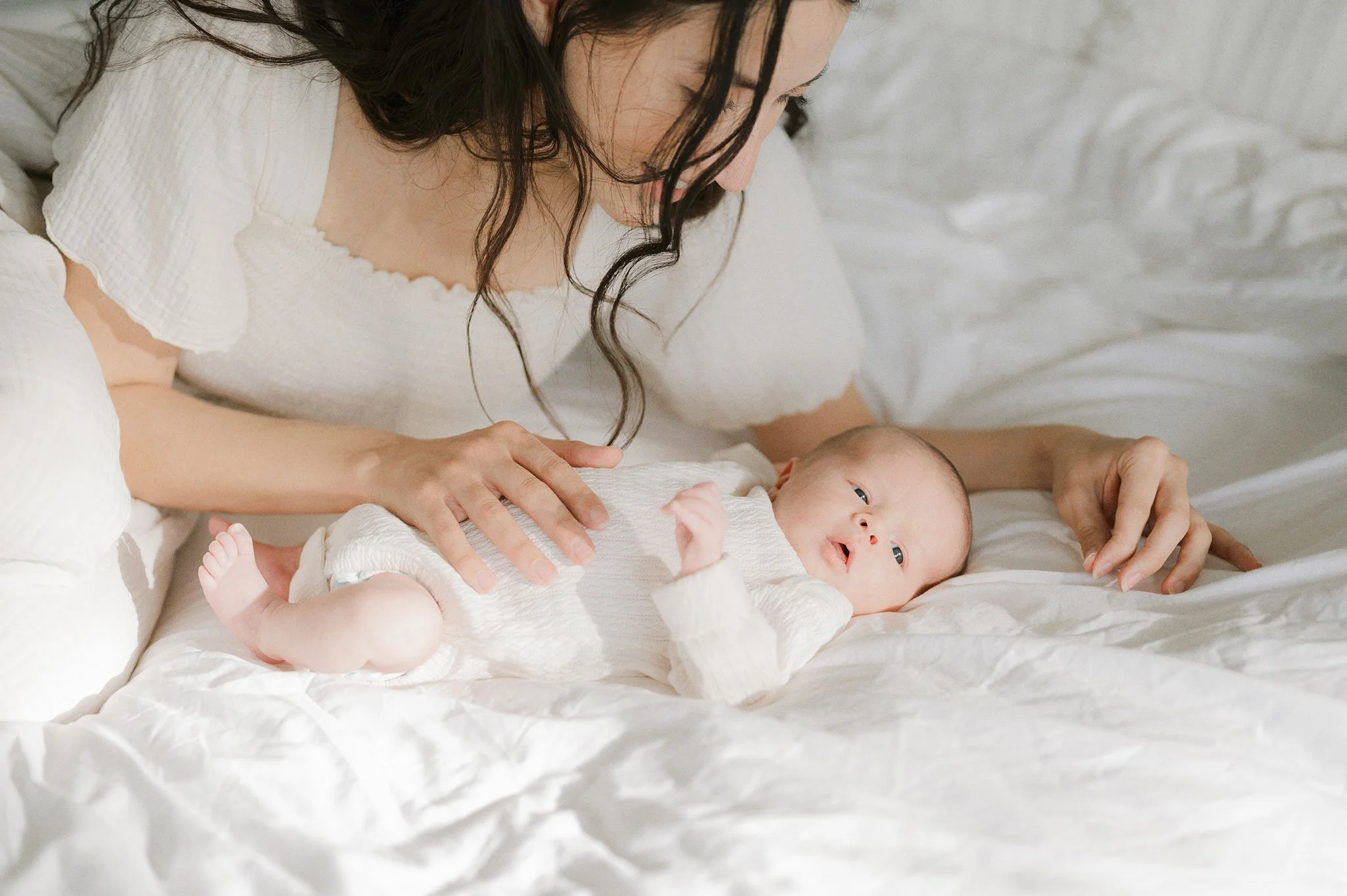 Mother with baby on a bed, both in white clothing, in a peaceful setting.