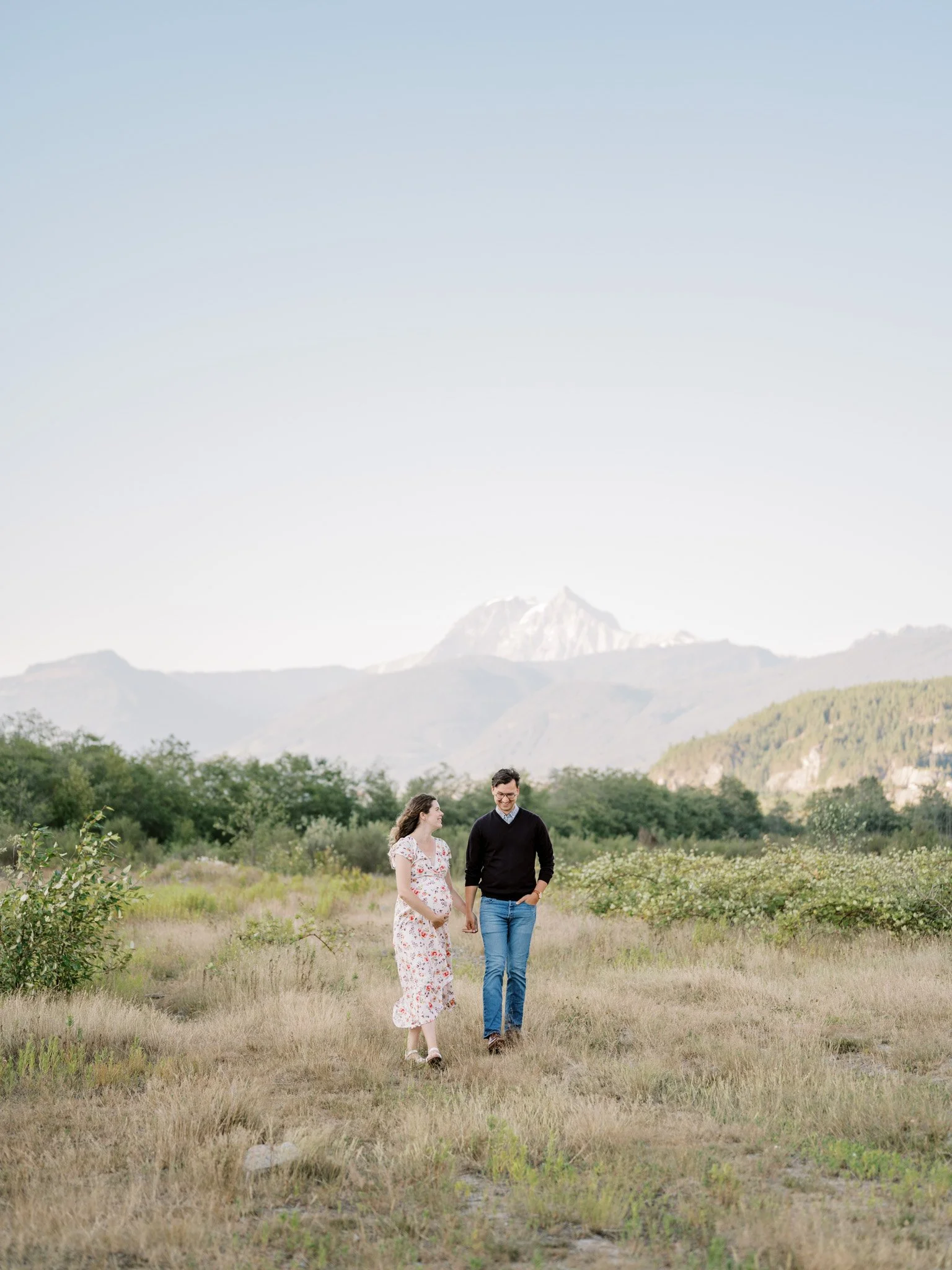 A couple walking hand in hand through a grassy field with mountains in the background.