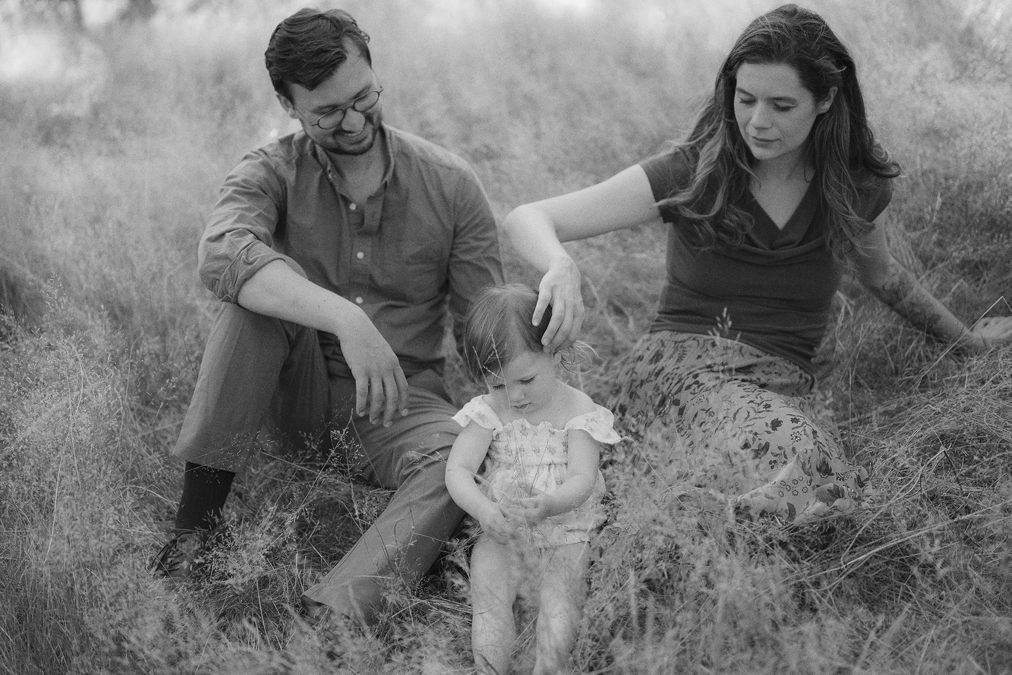 A family of three sitting on a grassy field, with a man and woman looking down at a young girl who is playing with the grass.