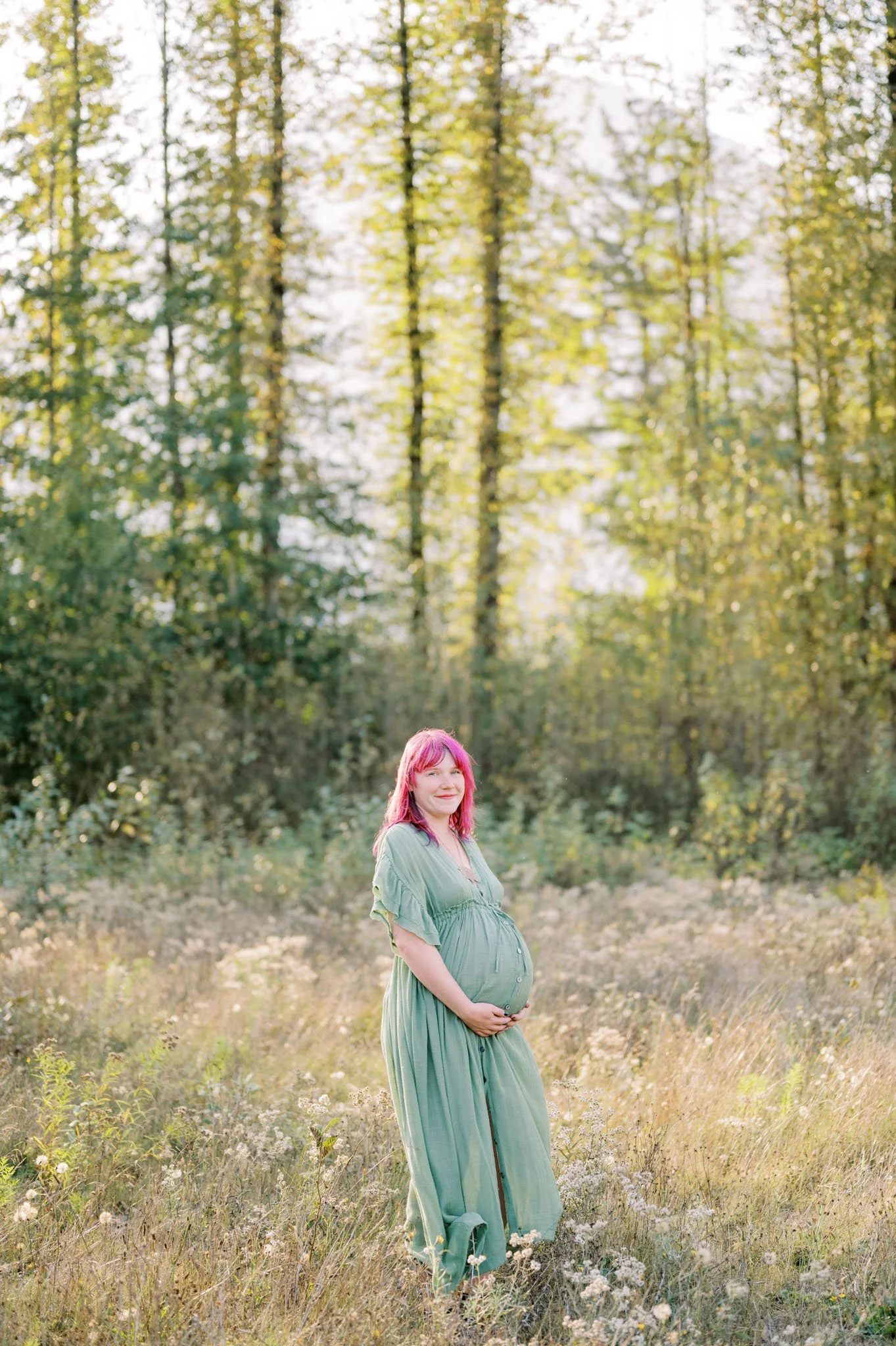 Pregnant woman with pink hair in a green dress standing in a sunny meadow with tall trees in the background.