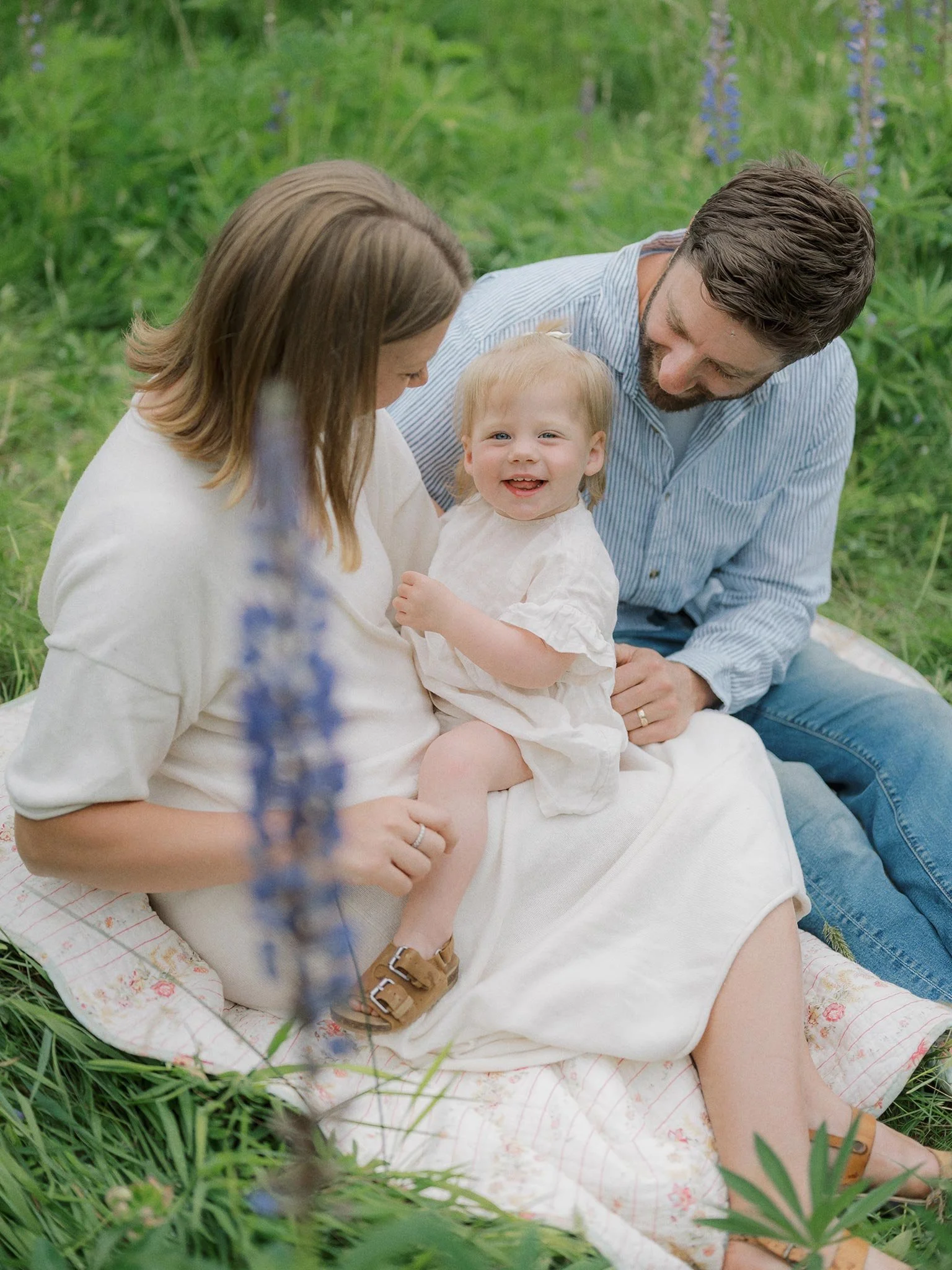 A happy family of three sitting on a blanket in a green outdoor setting, with a young child smiling.