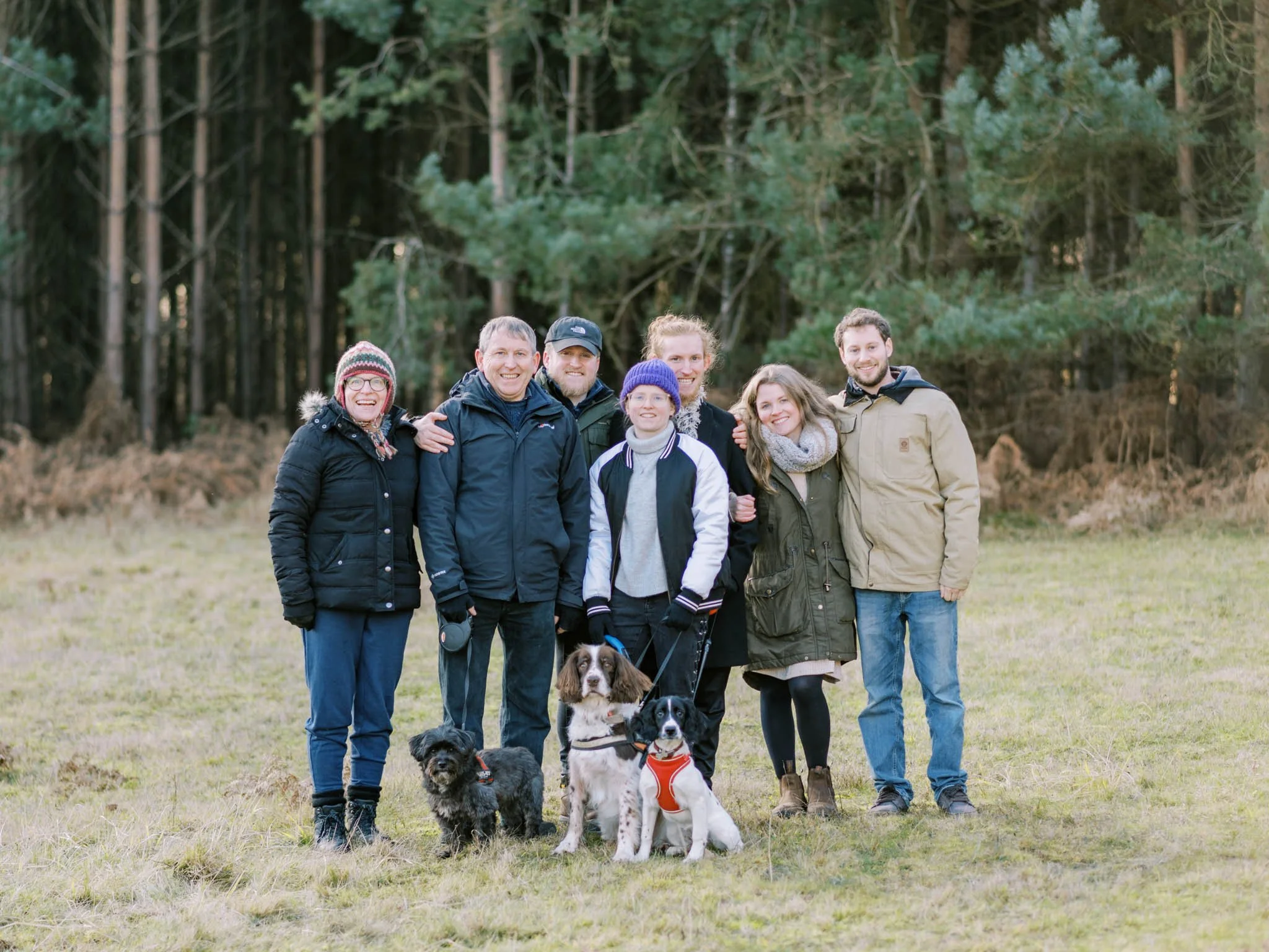 Group of people and dogs in a field with pine trees in the background.