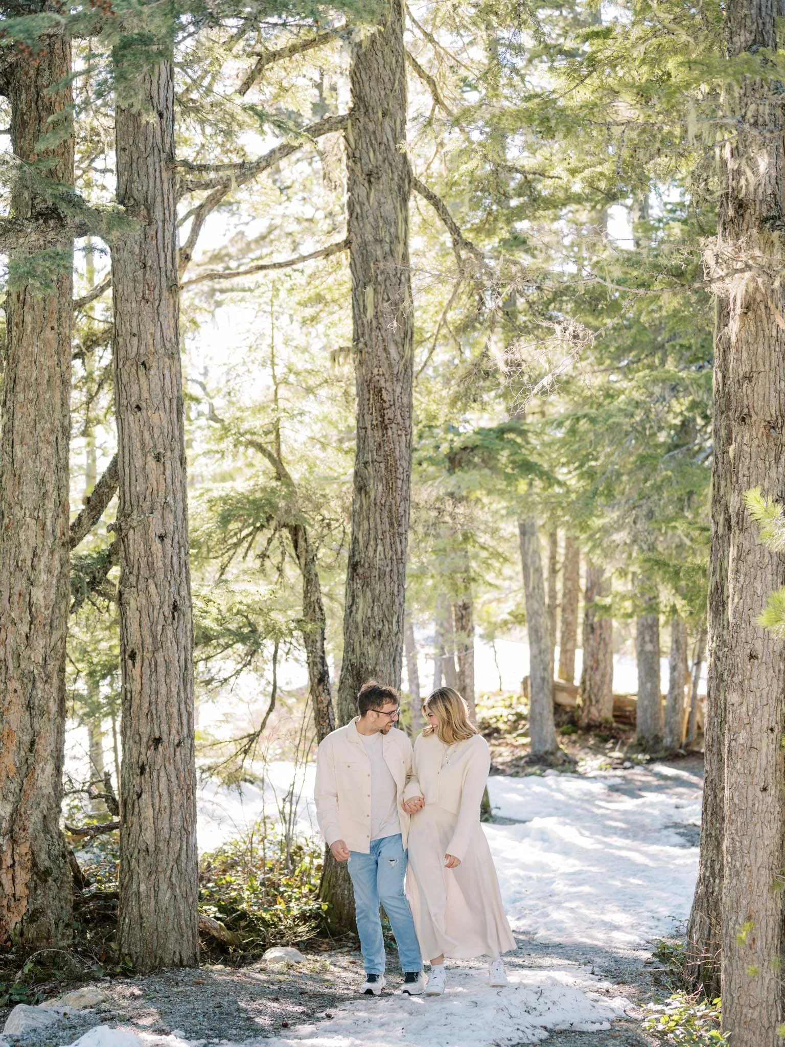 Couple walking in a snow-covered forest with sunlight filtering through tall trees.