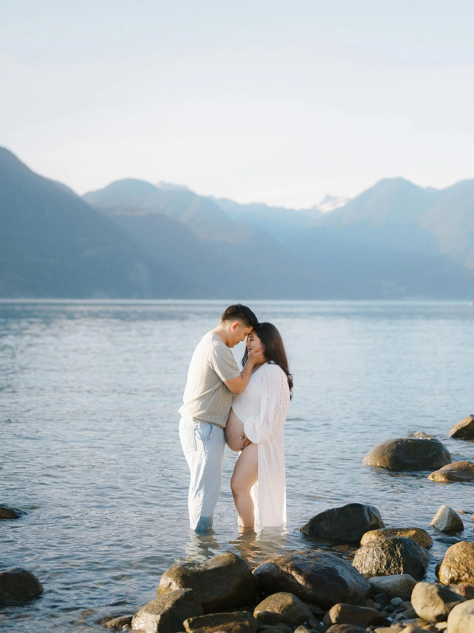 A pregnant woman and a man standing in shallow water by rocks in a lake, embracing and touching foreheads with mountains in the background.