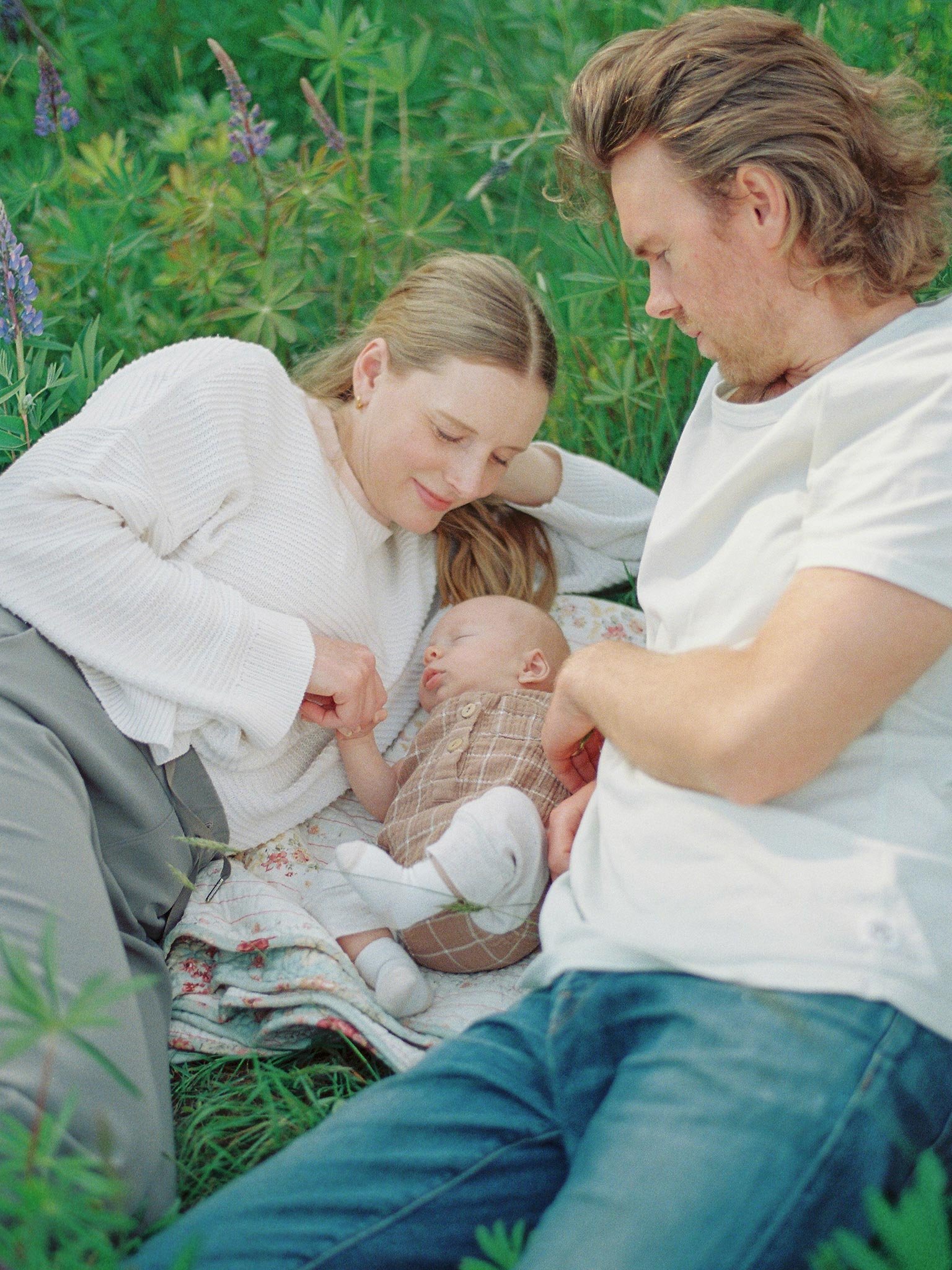 A young woman and a man, likely her partner, are laying on a patch of grass with a newborn baby. The woman is gently holding the baby's hand and looking at him affectionately, while the man looks on. The scene is outdoors with lush green foliage in the background.