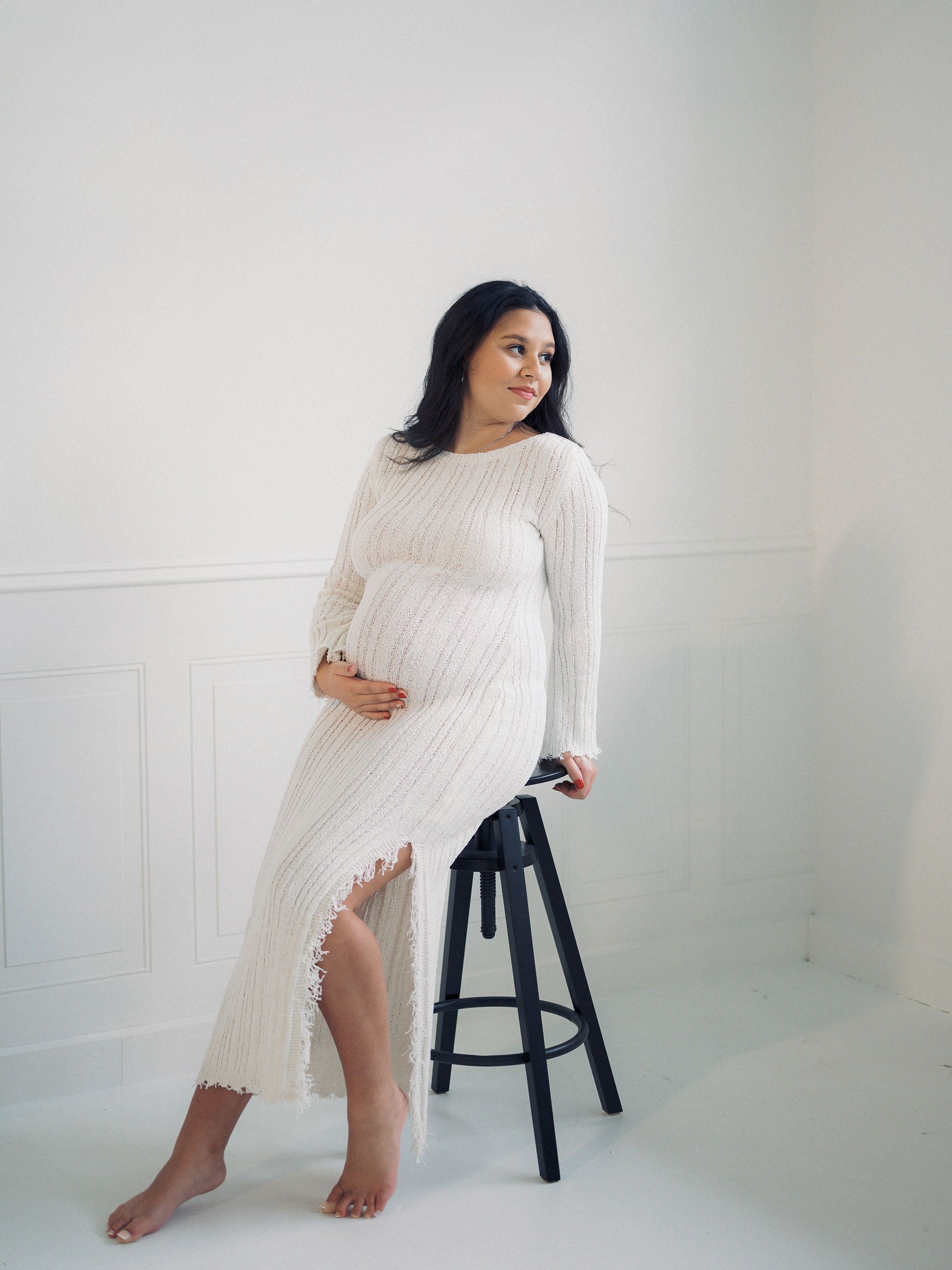 Pregnant woman in white dress sitting on a stool touching her belly.