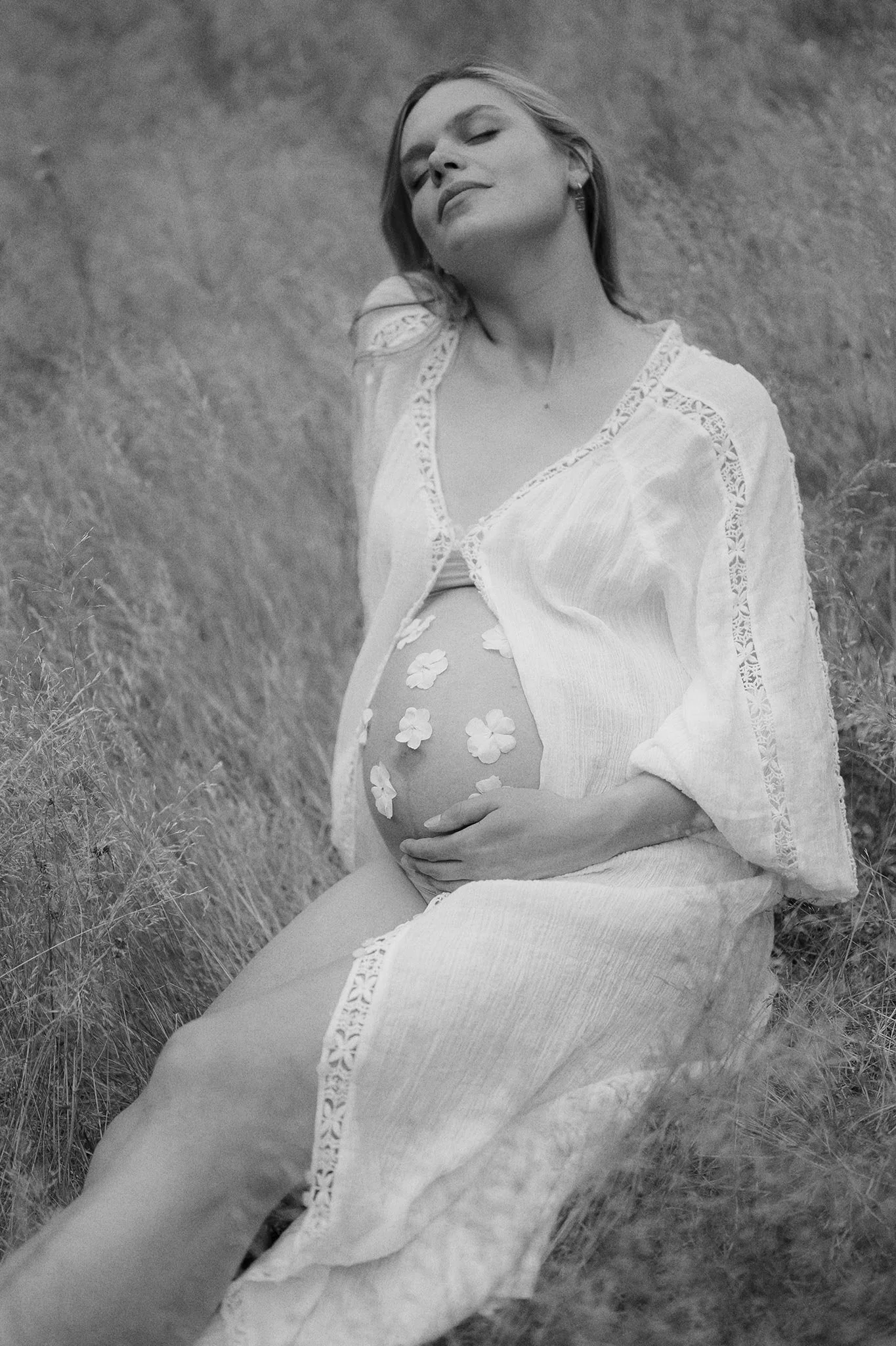 A pregnant woman sitting outdoors in tall grass, wearing a light-colored dress and an open cardigan, with flowers on her belly, smiling softly.