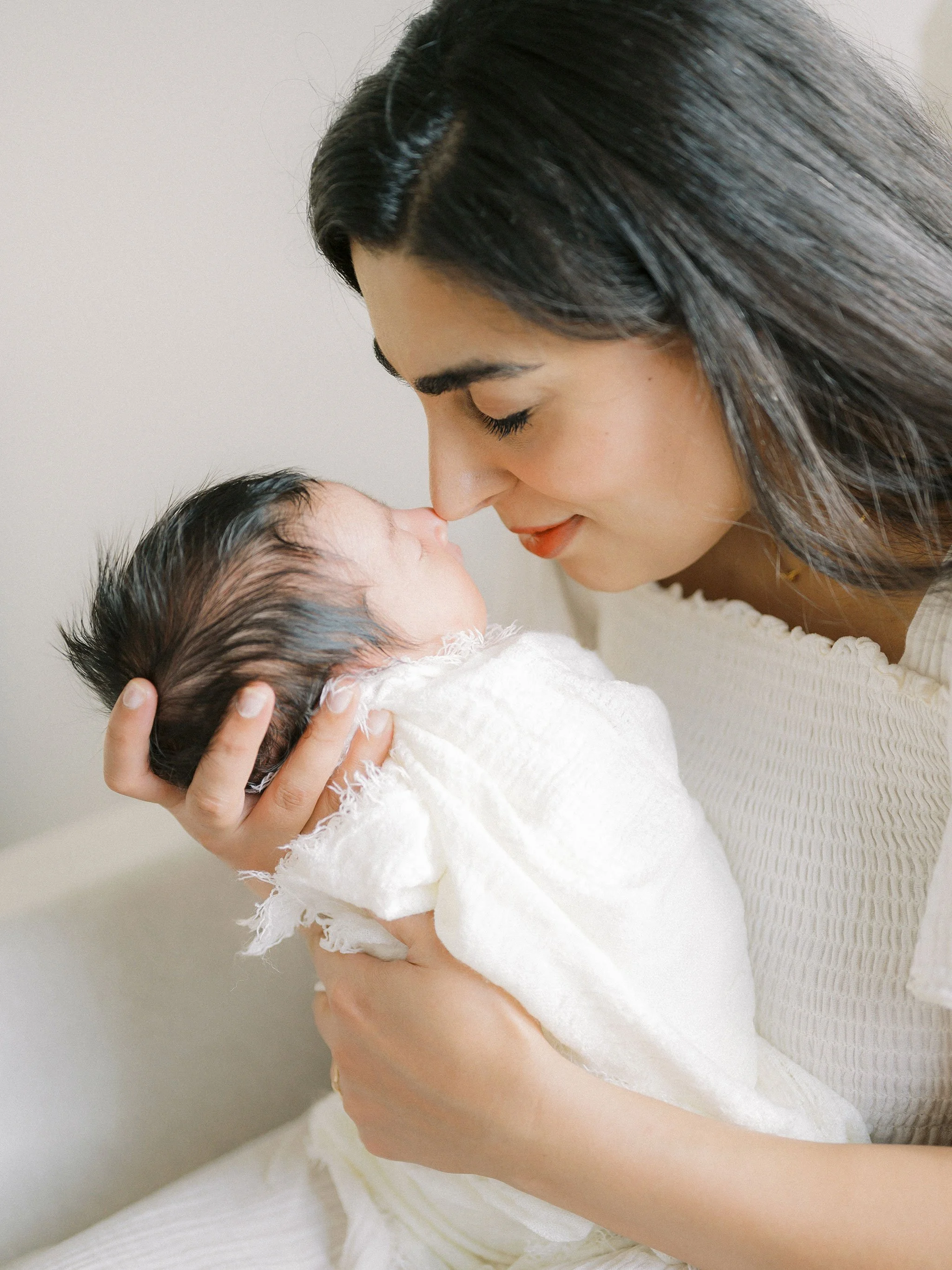 A woman gently holding a newborn baby wrapped in a white blanket, touching noses with the baby.