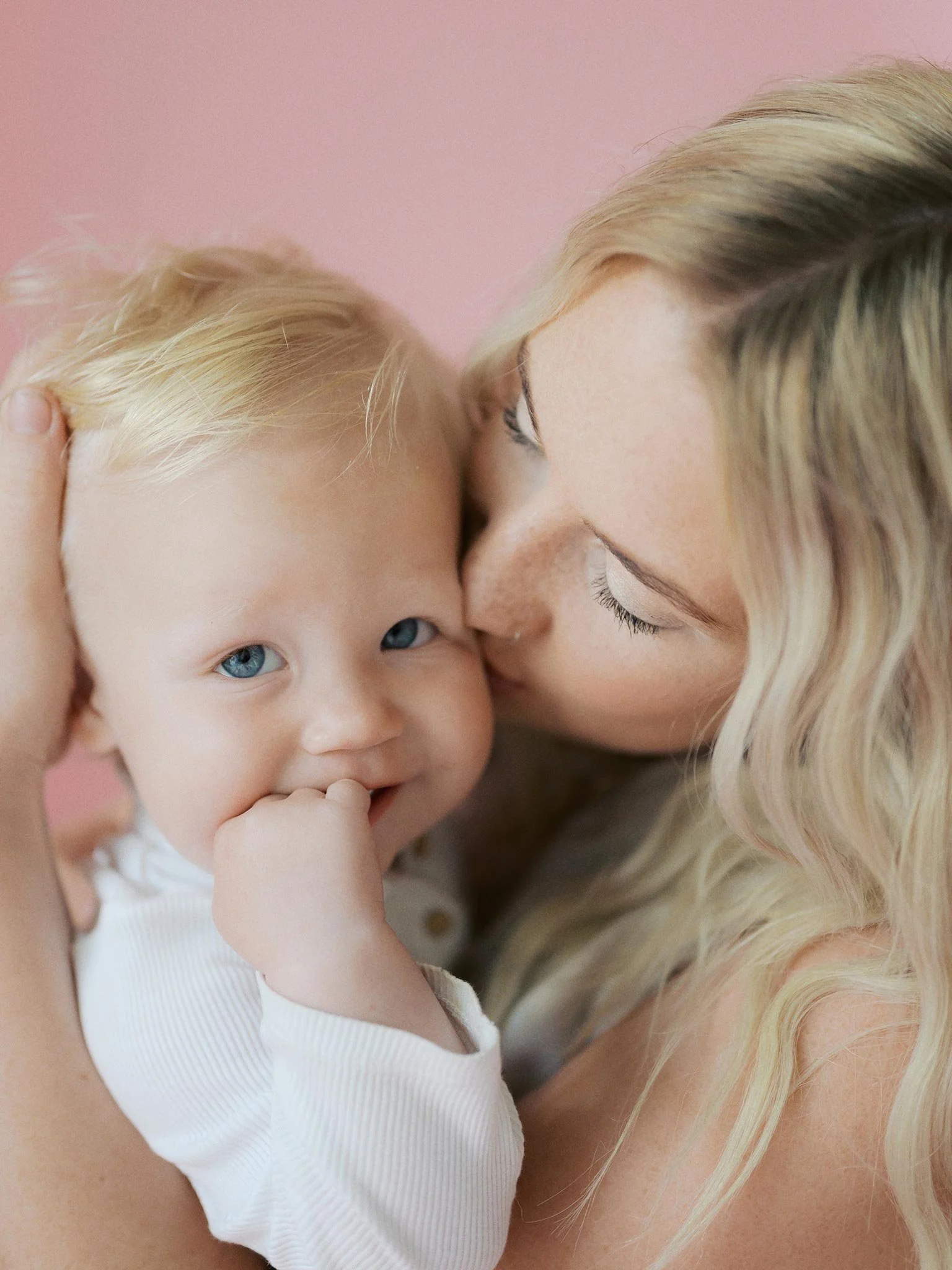 A woman with blonde hair kissing a toddler with blonde hair and blue eyes, who is smiling and sucking on fingers, against a pink background.