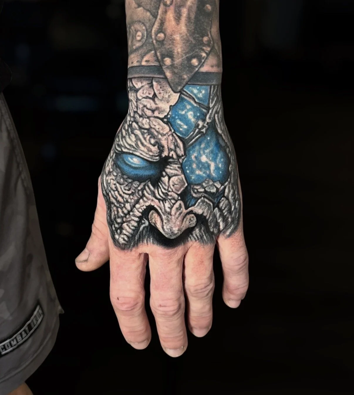 Black and grey cracked skull hand tattoo with bright blue eye and cosmic accents, by Chris Kennedy.
