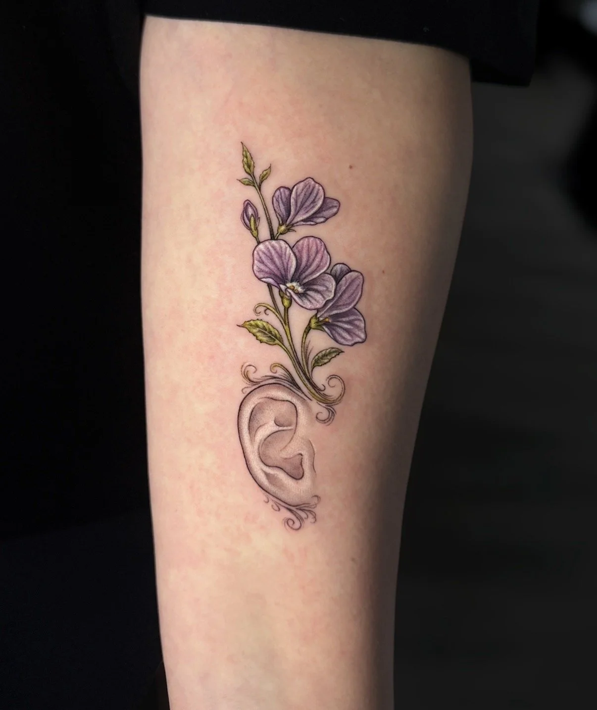 Purple flower tattoo growing above a softly shaded ear with decorative stems and leaves, by Chris Kennedy.