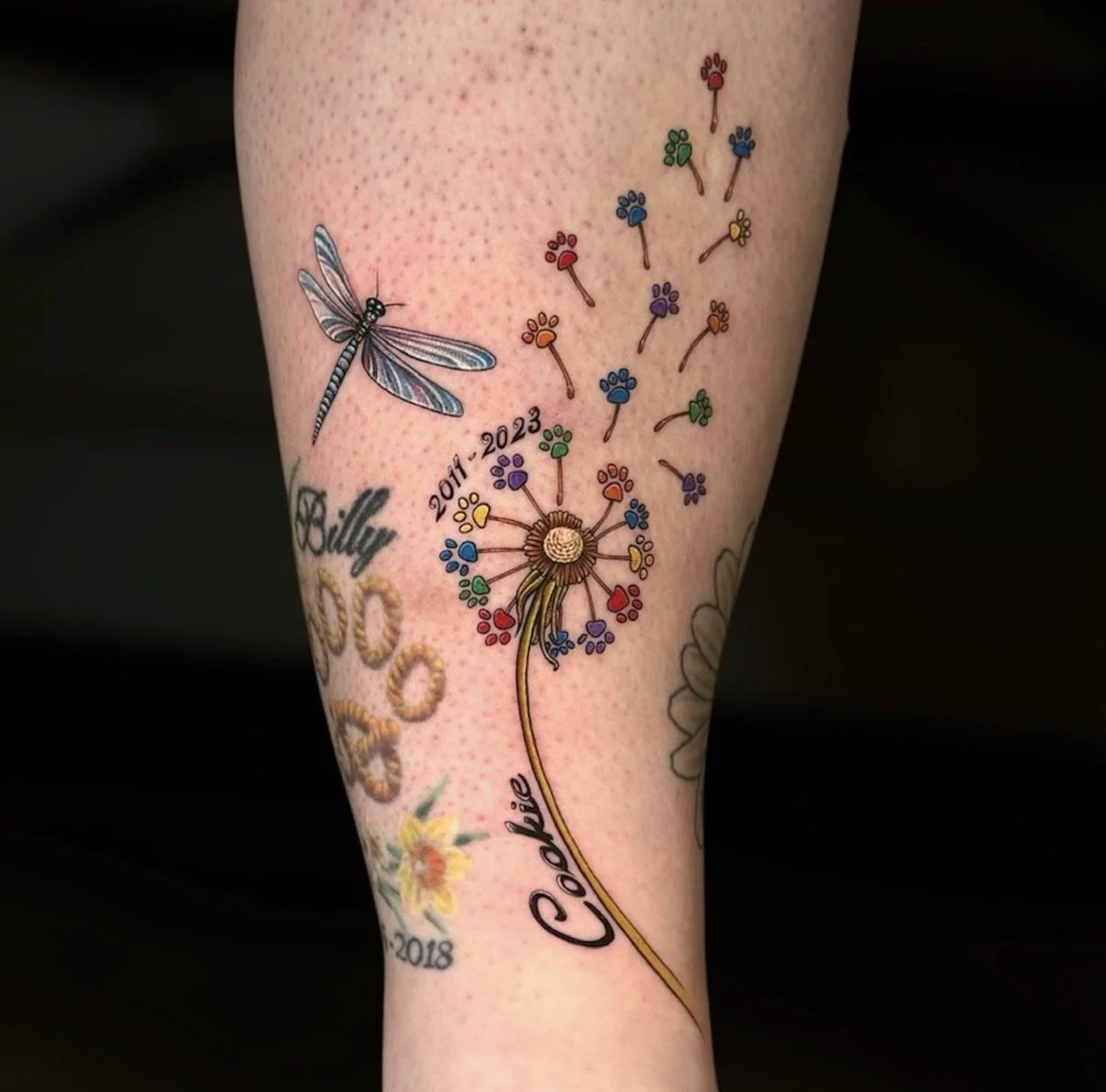 Colour memorial dandelion tattoo with rainbow paw-print seeds, a dragonfly, the name Cookie and dates 2011–2023, by Chris Kennedy.