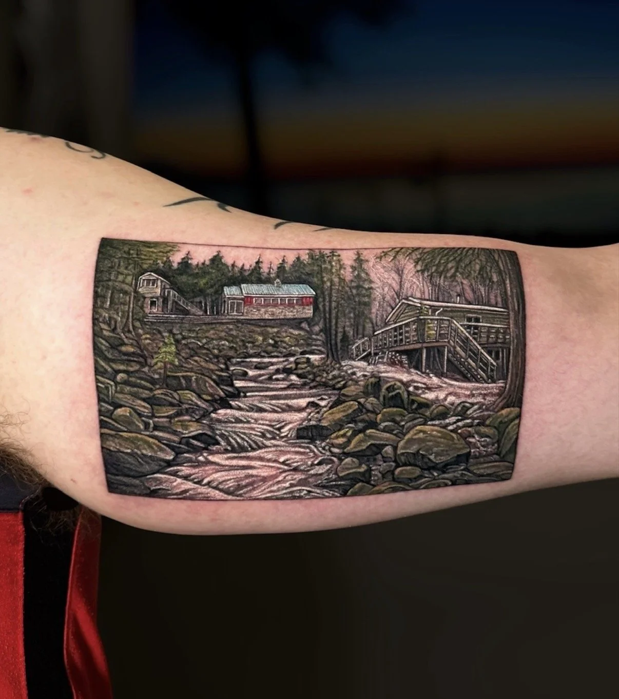 Colour landscape tattoo of a rocky forest stream with cabins and a red railway carriage, by Chris Kennedy.