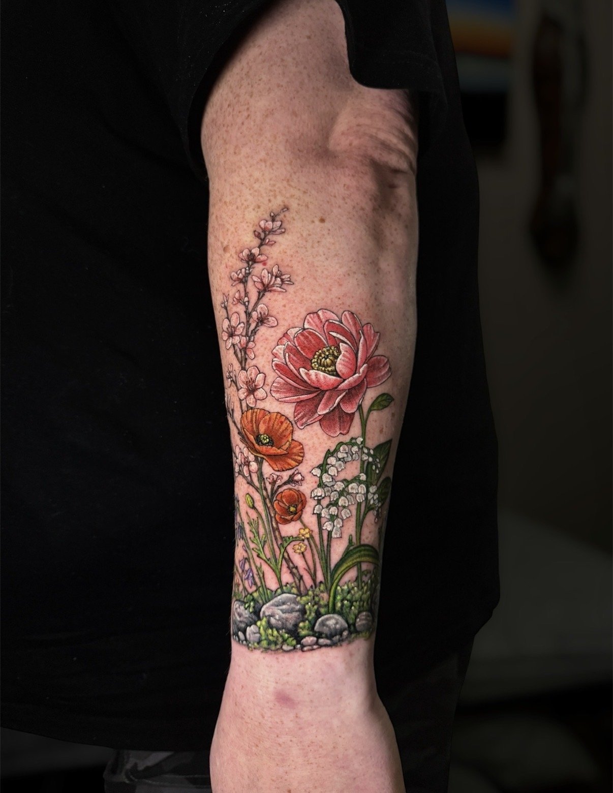 Colour garden tattoo with pink and orange flowers, lily of the valley, green foliage and small stones, by Chris Kennedy.