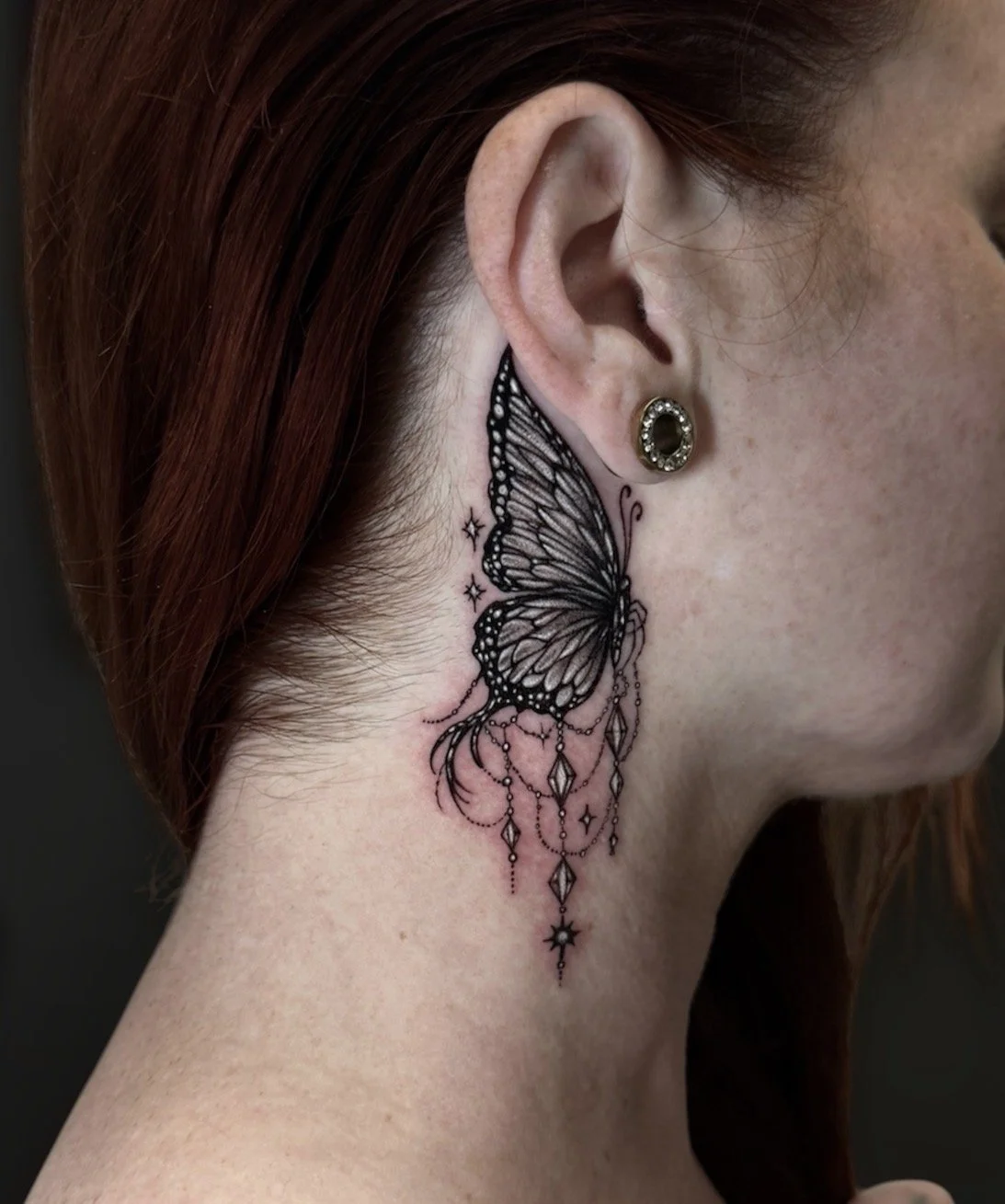 Black and grey butterfly tattoo behind the ear with dangling gem ornaments and small stars, by Chris Kennedy.