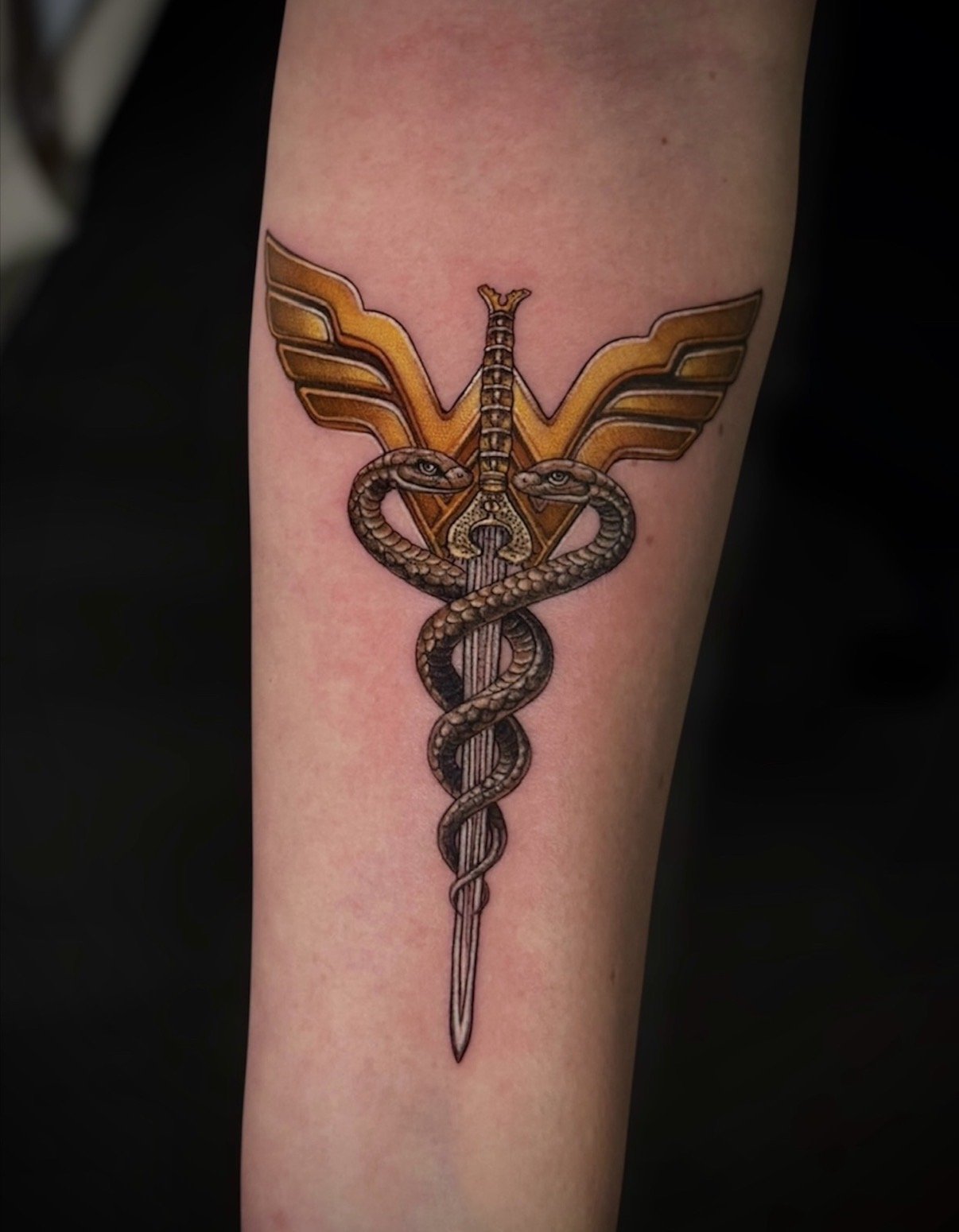 Colour Wonder Woman emblem tattoo with two snakes wrapped around a sword, by Chris Kennedy.