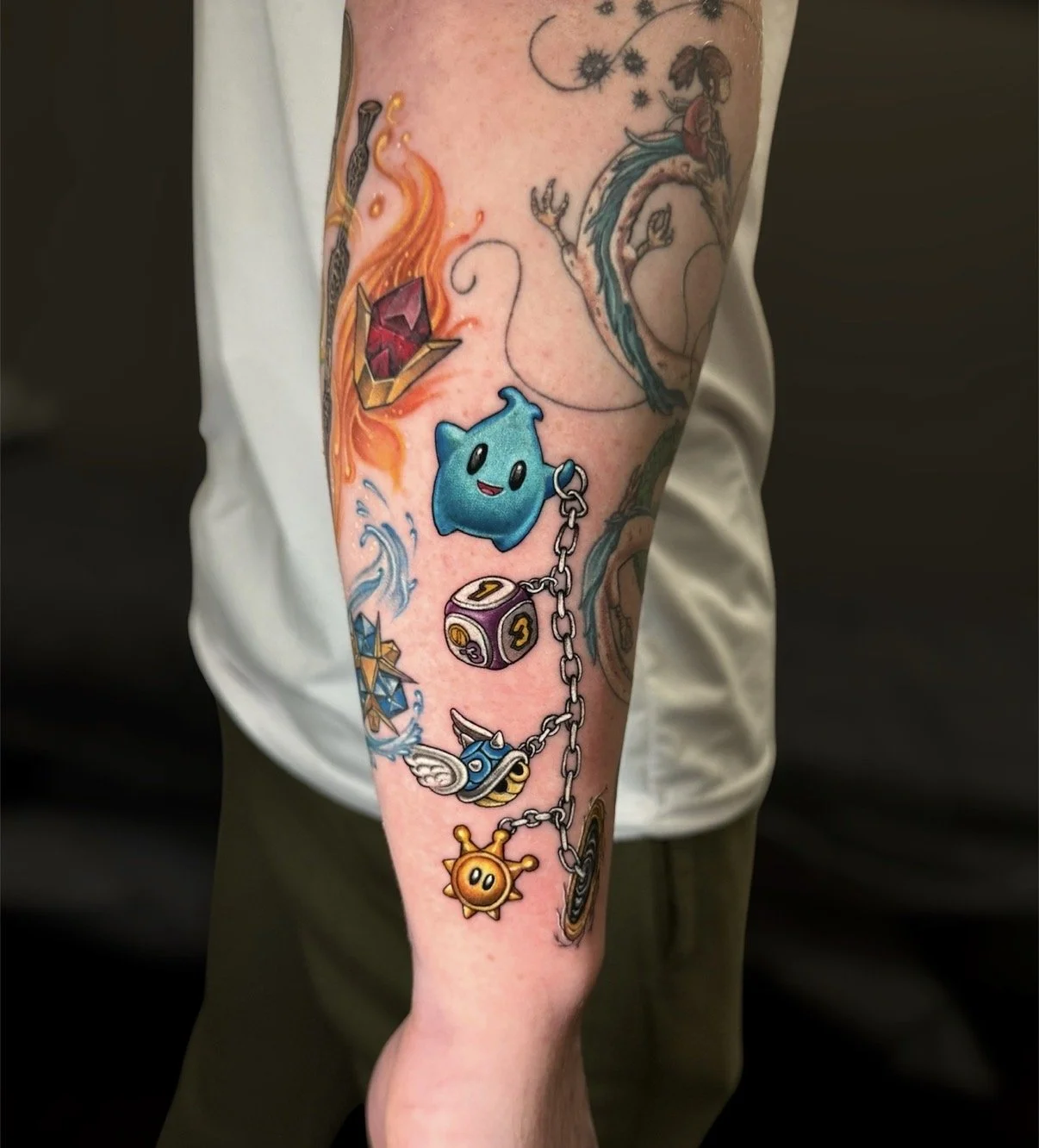 Colour gaming charm tattoo with a blue star character, dice block, winged blue shell and golden sun linked by a chain, by Chris Kennedy.