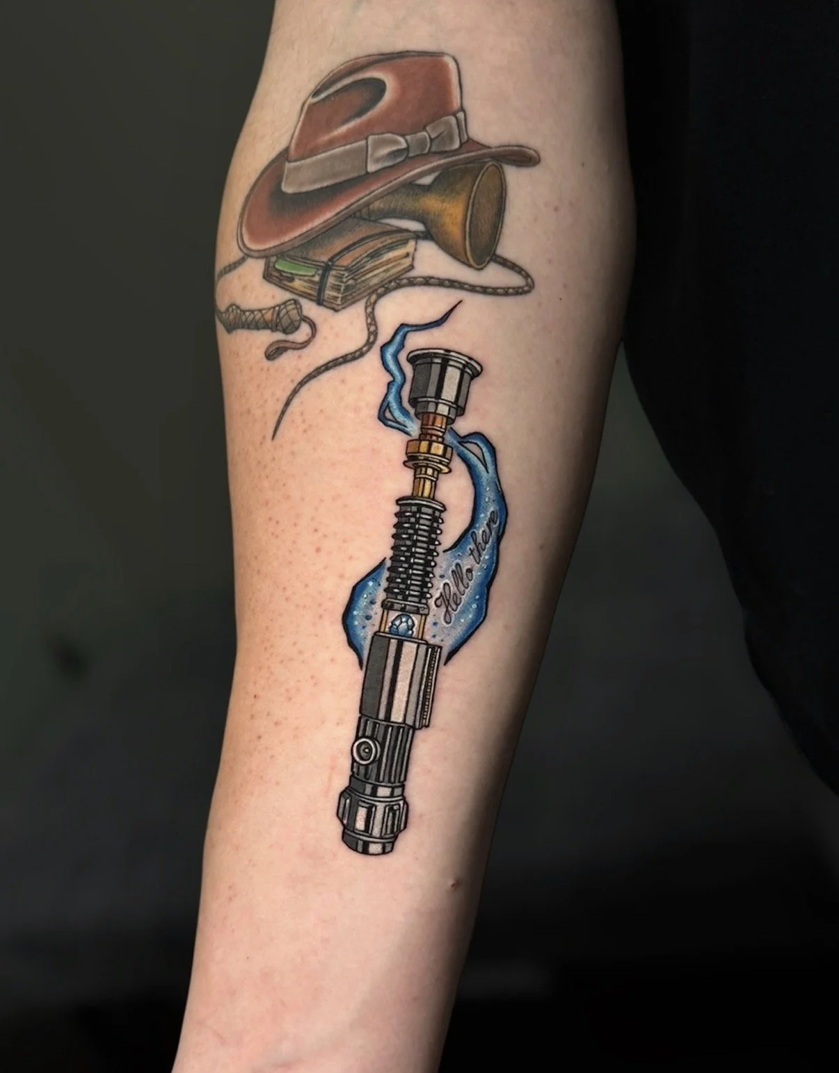 Colour lightsaber tattoo with blue accents beneath an Indiana Jones hat, by Chris Kennedy.