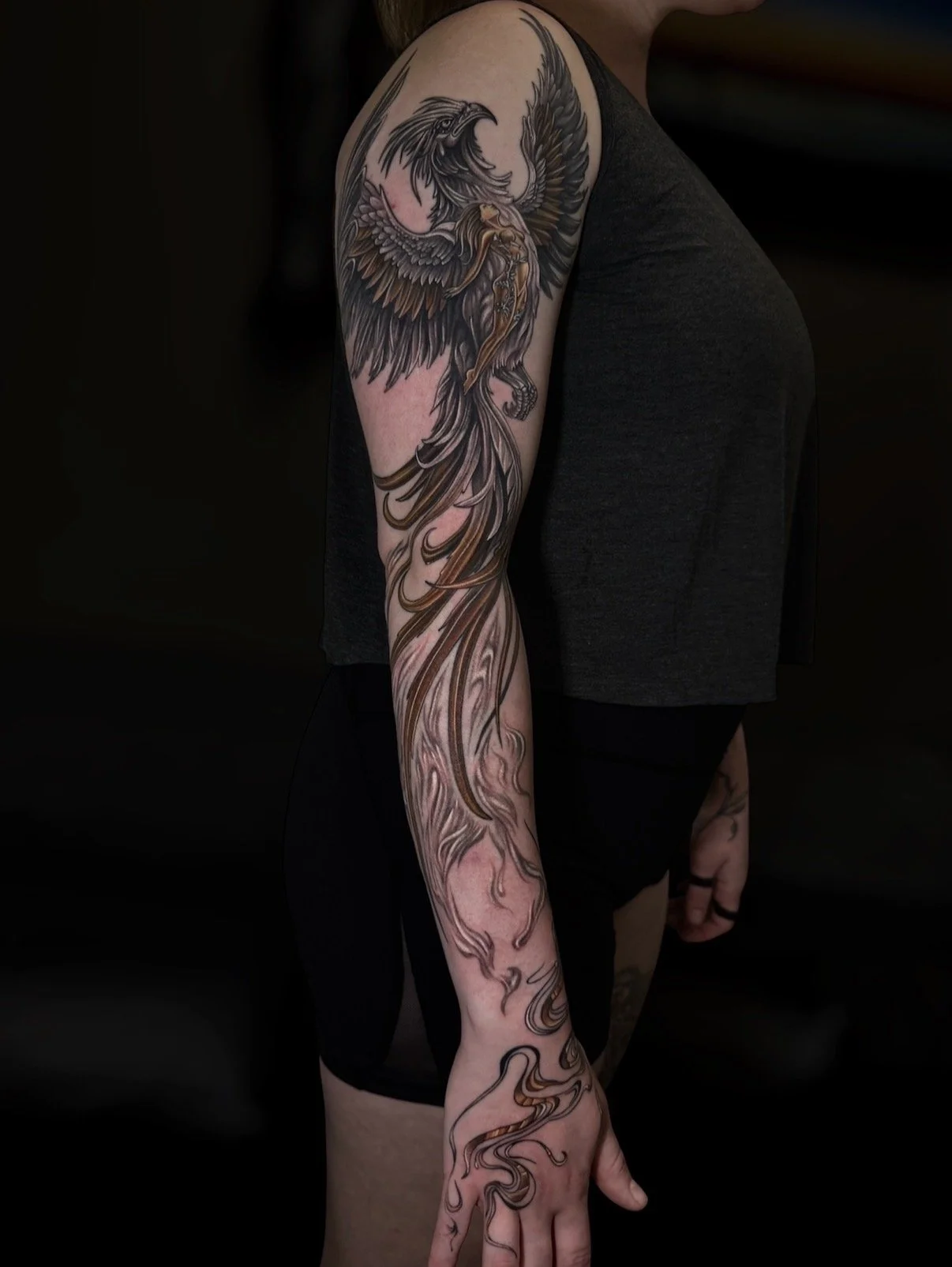 Black and grey phoenix sleeve tattoo with a female figure and warm gold feather accents extending onto the hand, by Chris Kennedy.