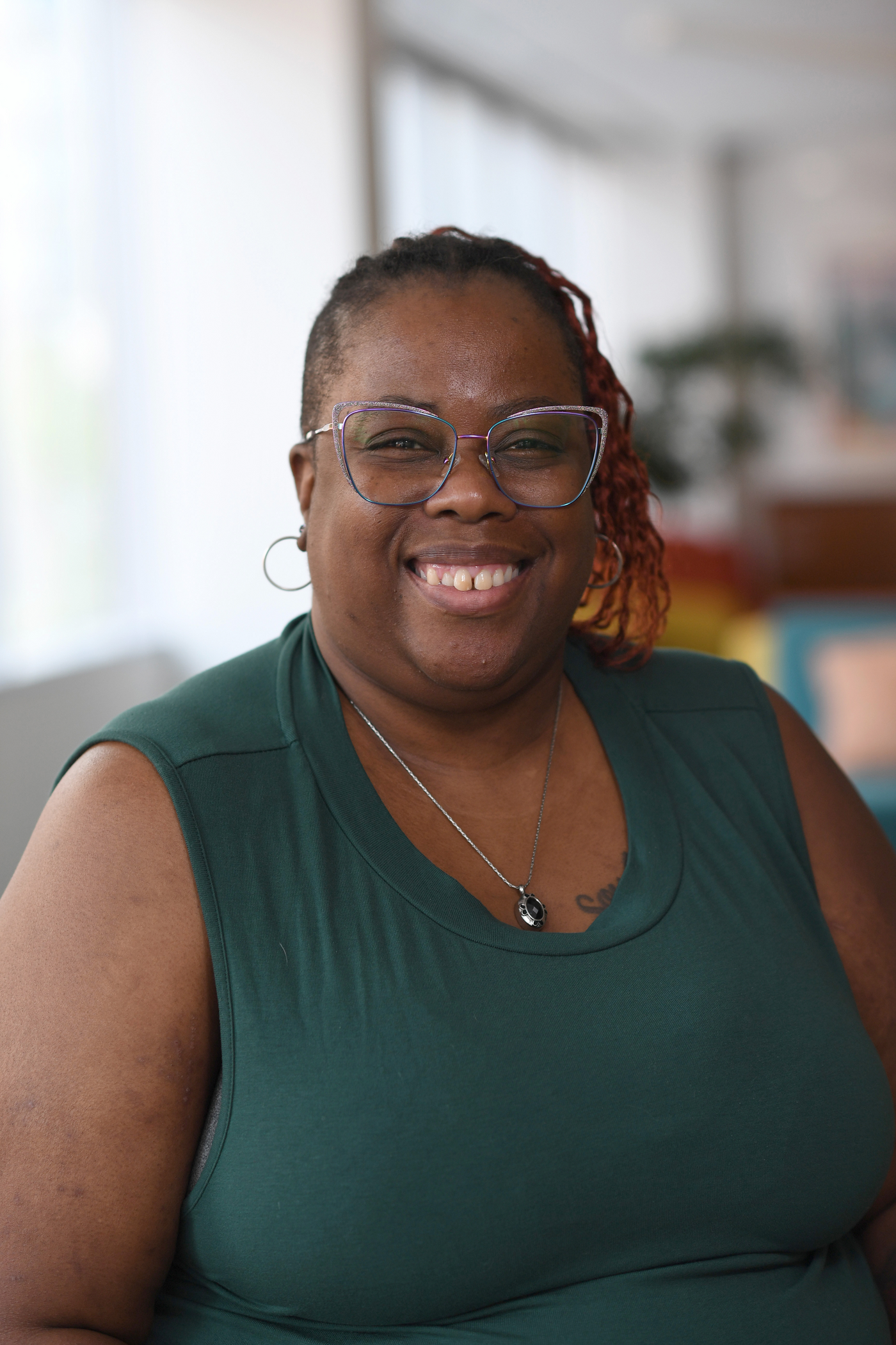 miling woman in glasses and green top seated in a bright, welcoming room.