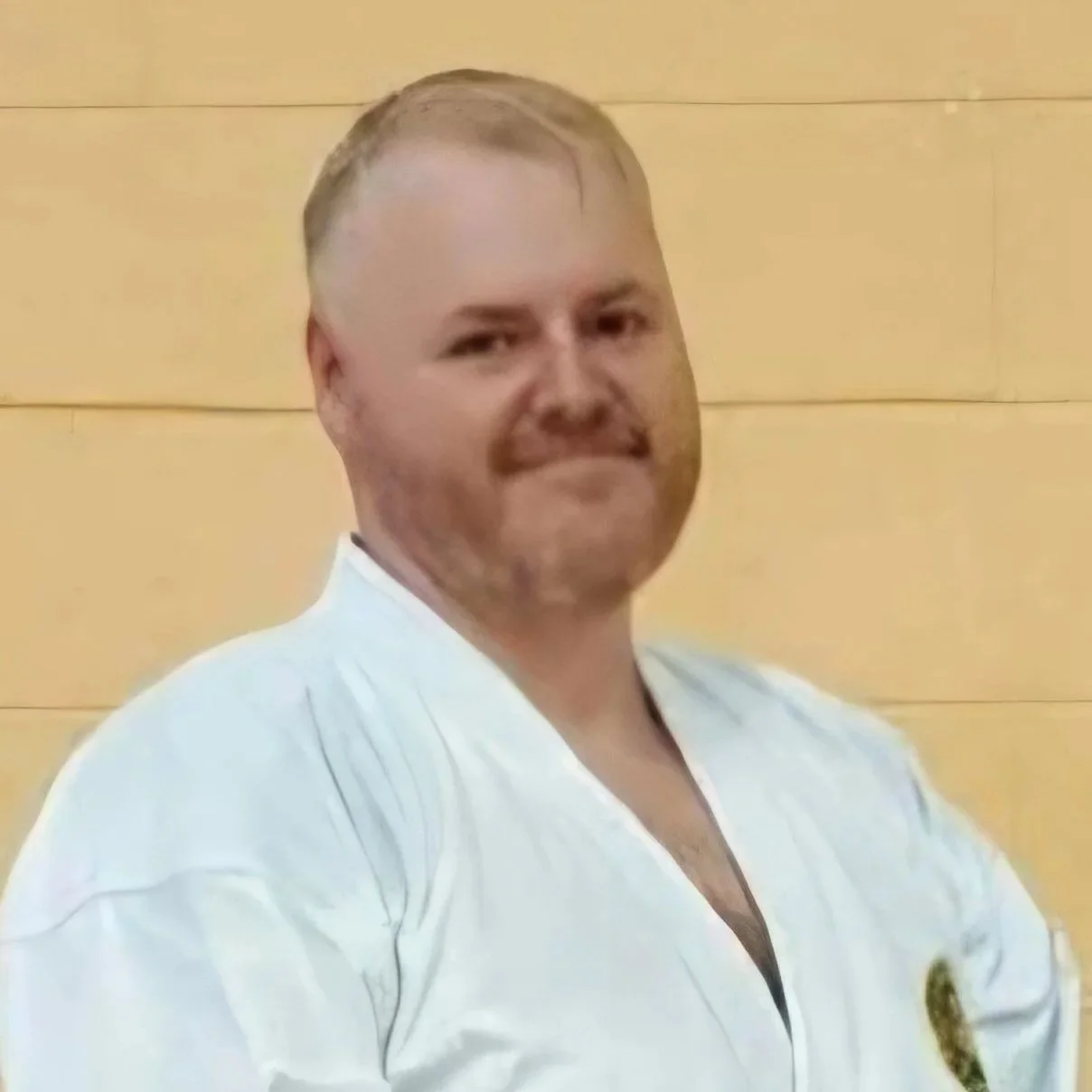 A man with short, light-colored hair and a goatee, wearing a white martial arts uniform, standing in front of a wooden wall.