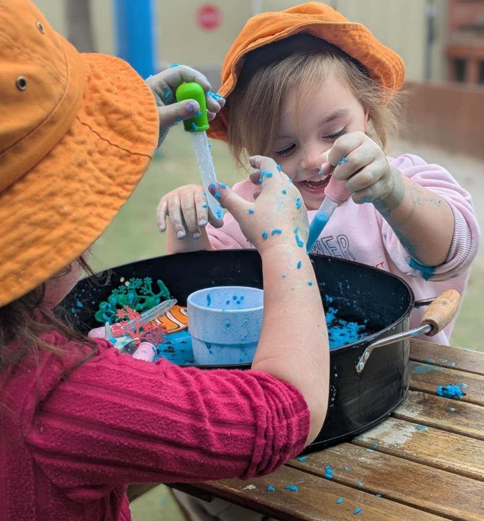 Two young children, one with a pink shirt and the other with an orange hat, are playing with blue slime and toys in a black tray outdoors. They are smiling and splattered with the slime.